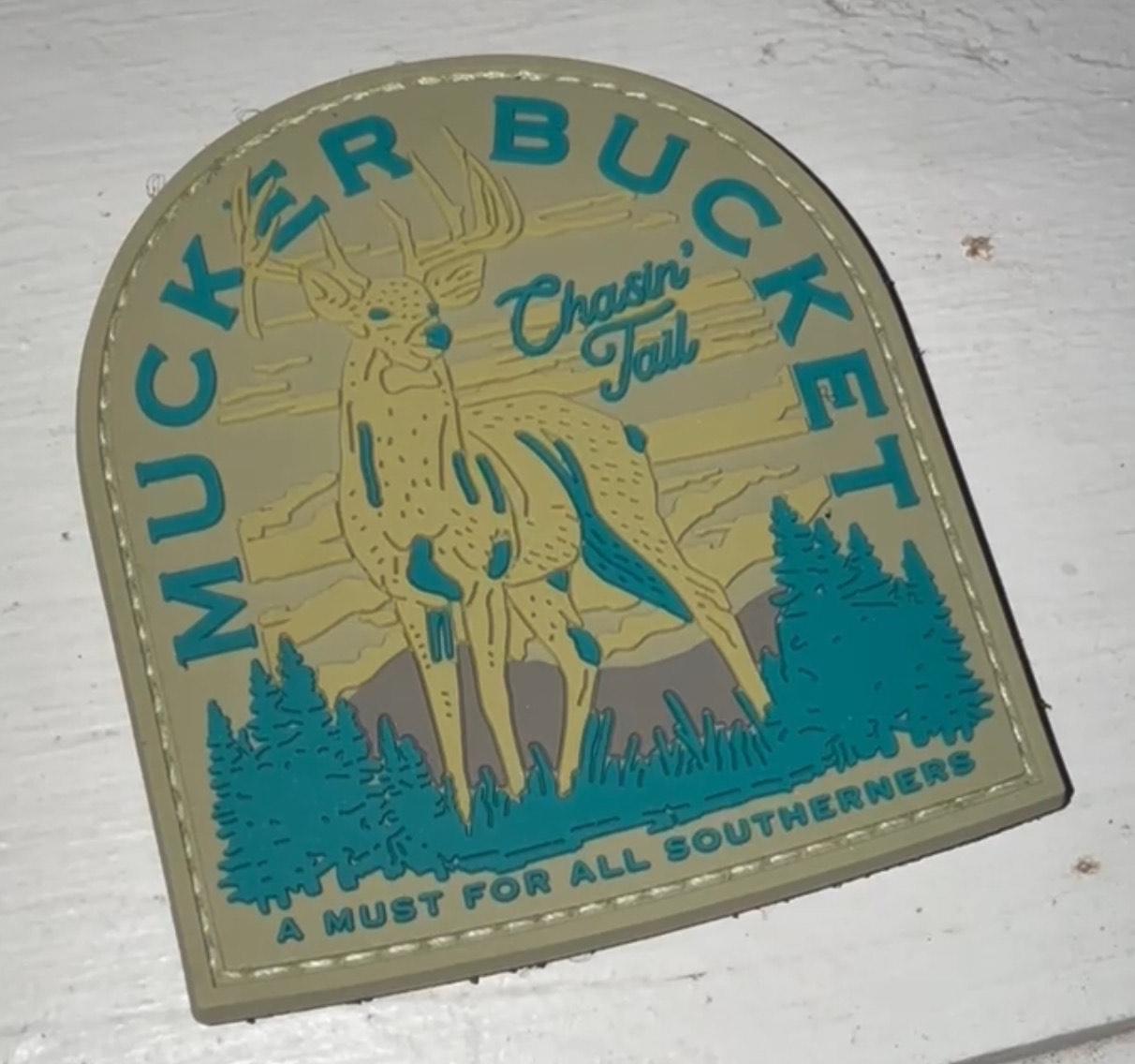 The Chasin Tail 2.0 PVC Patch by Mucker Bucket features a deer, pine trees, and mountain design with MUCKER BUCKET and Chasin Tail text. Its beige, teal, and yellow tones make it essential for Southerners on the hunt.
