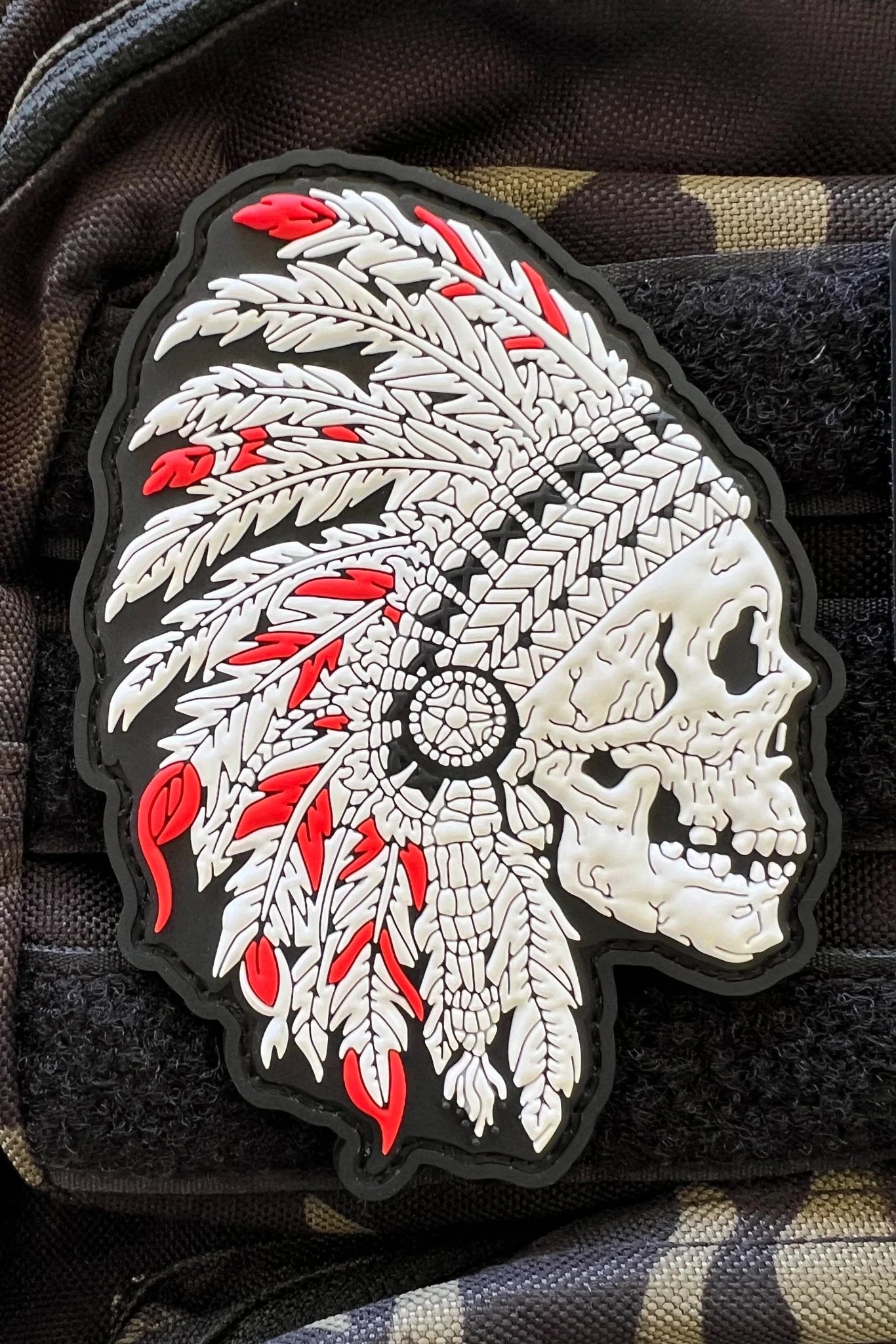The Trustless Tribe PVC Patch from Relentless Betrayal displays a 3D skull with a feathered headdress. Its white feathers outlined in black and striking red accents stand out on any dark fabric, whether its on a bag or clothing.