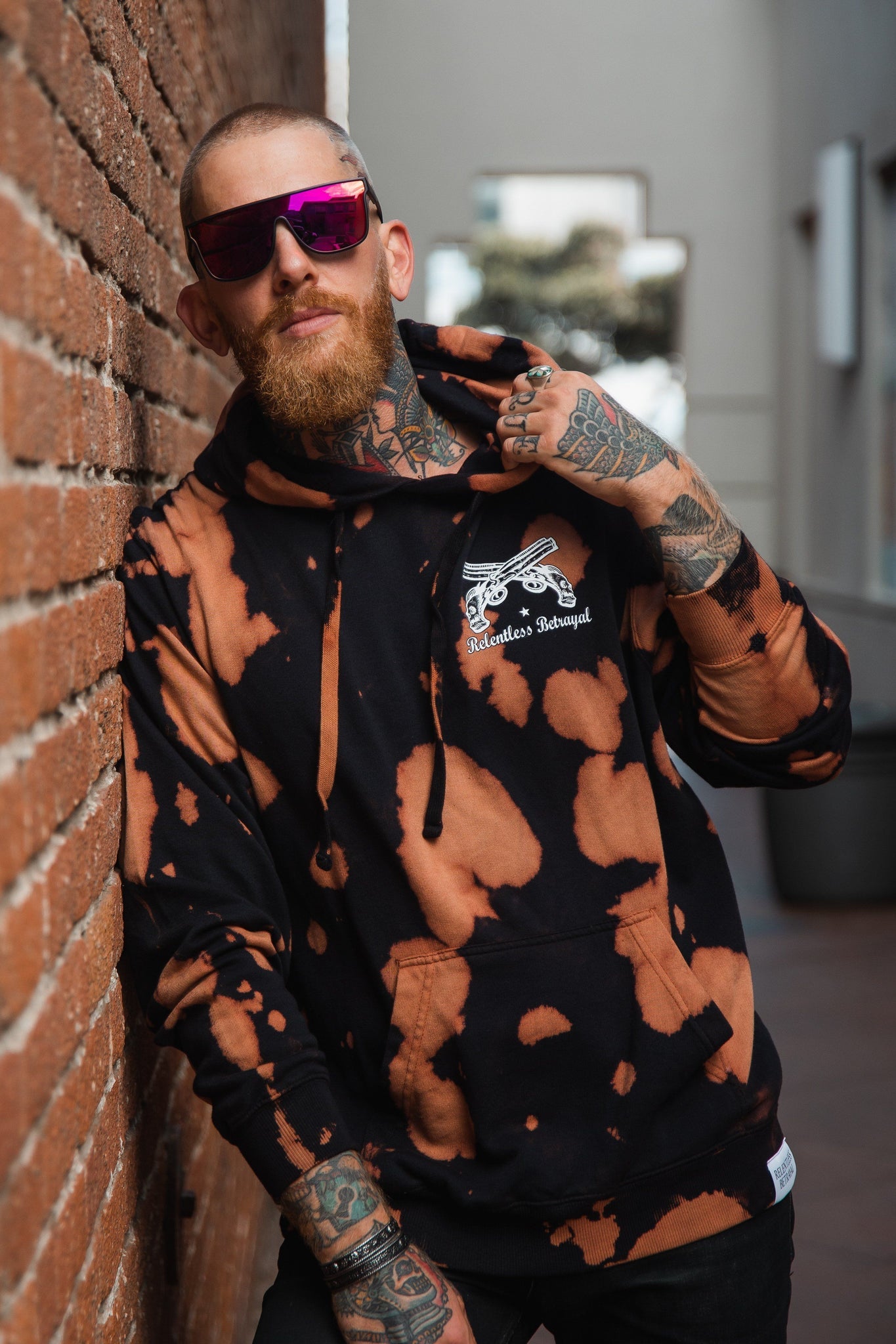 A bearded man with tattoos leans against a brick wall, wearing the No Prey, No Pay Bleach Wash Hoodie by Relentless Betrayal. With its cotton polyester blend and logo with crossed bones design, it perfectly complements the urban vibe of the outdoor corridor.