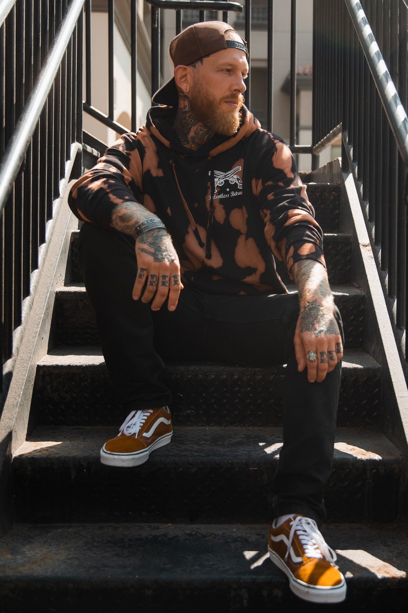 A tattooed person sits on an outdoor staircase, wearing a No Prey, No Pay Bleach Wash Hoodie by Relentless Betrayal made from cozy cotton polyester blend fleece. Theyre in dark pants and brown sneakers, have a beard, wear a backward cap, and gaze off to the side.