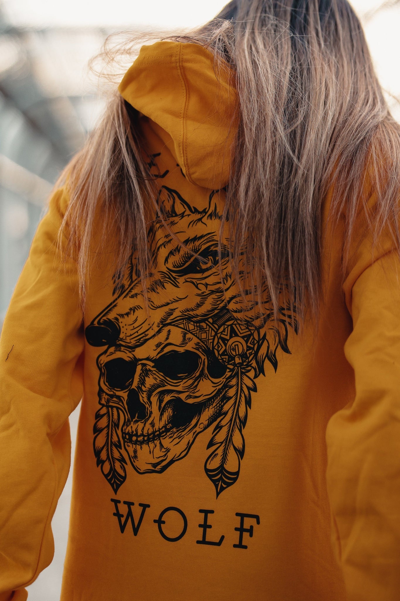 A person with long hair dons the Lone Wolf GOLD Hoodie by Relentless Betrayal, showcasing a black wolf head, skull, and feather design. WOLF is printed below the artwork on this unisex piece, symbolizing a lone wolf against a blurred background.