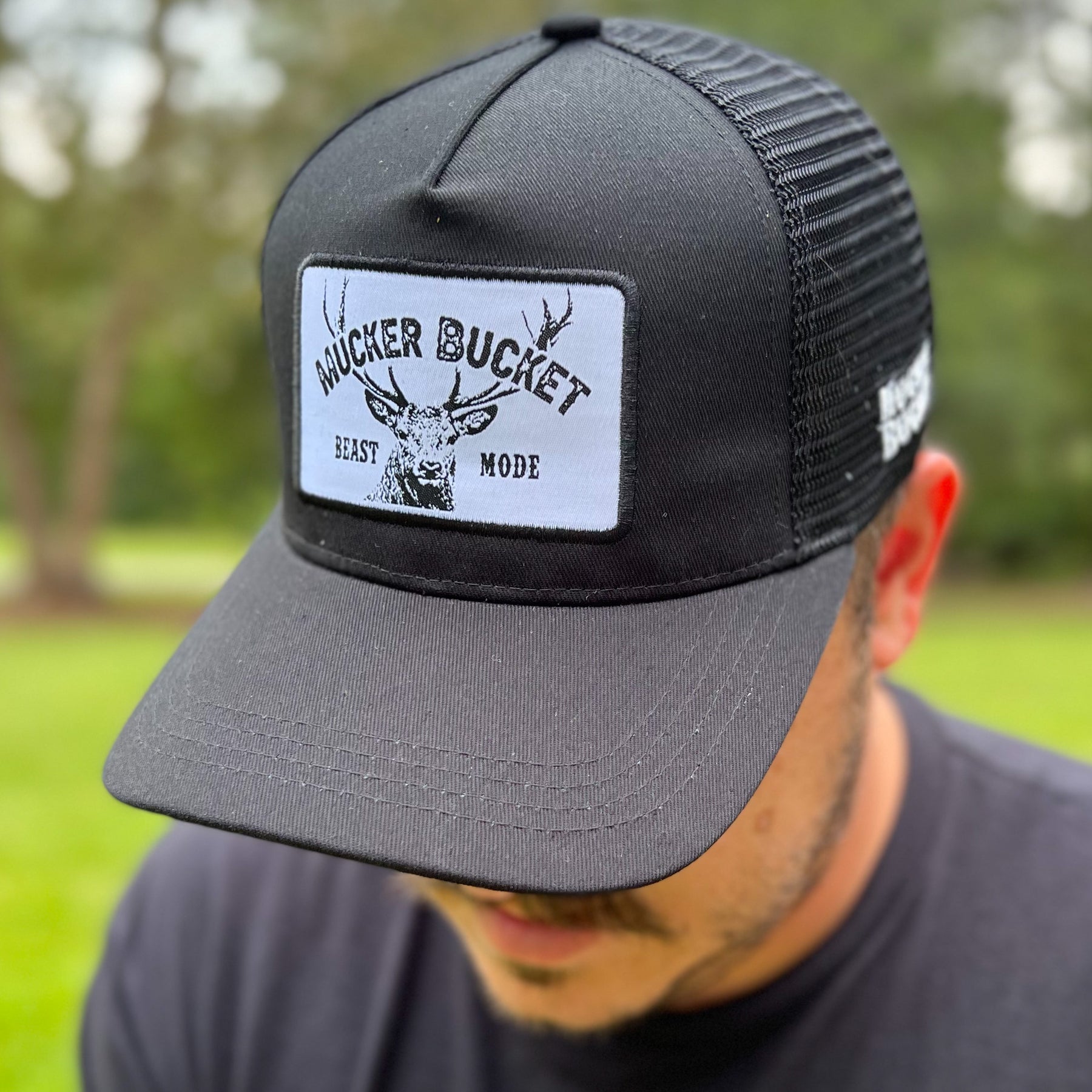 A person wears the Beast Mode OG Trucker Bucket by Mucker Bucket, a black snapback featuring a white-tail deer graphic and patch, against a blurred grassy and tree-filled backdrop.