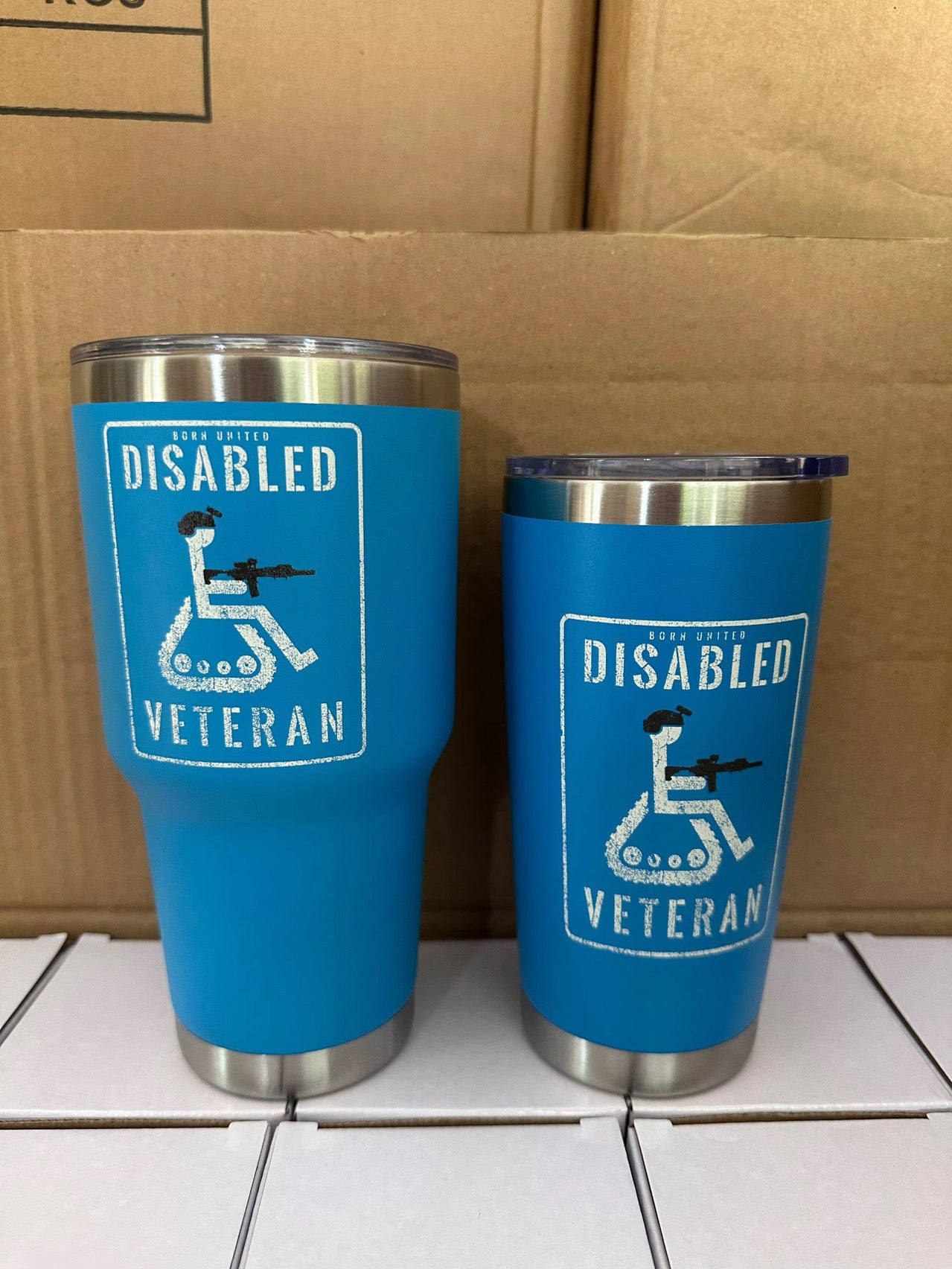 Two blue Born United Private Label Disabled Veteran Tumblers (30oz) with stainless steel bottoms sit side by side, each featuring a white logo of a wheelchair user holding a rifle and the words Disabled Veteran. Cardboard boxes are in the background.