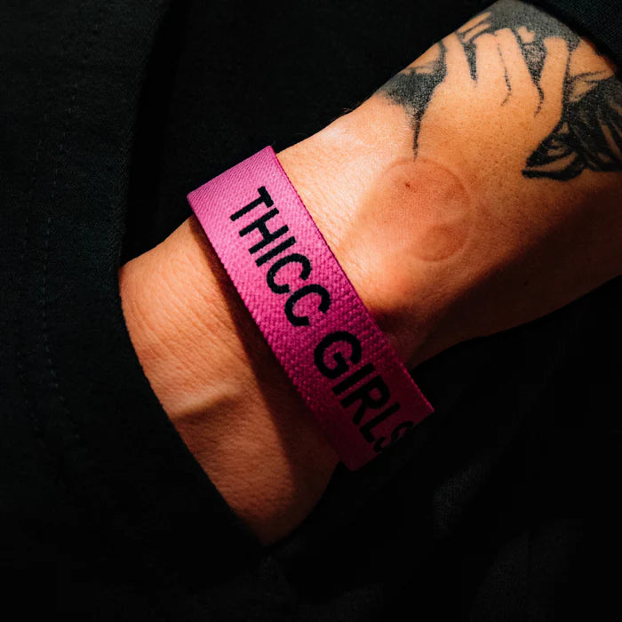 A tattooed wrist showcases the Thicc Girls Club Wristband by Tango Charlie Apparel, featuring bold black THICC GIRLS text on a pink band. Set against a dark backdrop, it celebrates body positivity with every glance.