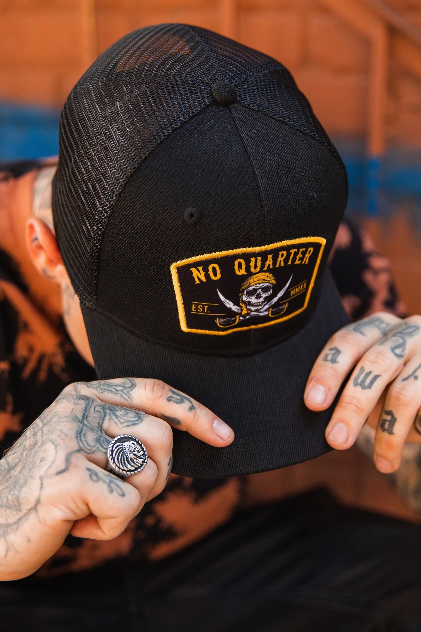 A person wearing a No Quarter Black Trucker Snapback by Relentless Betrayal, featuring a skull and crossed swords patch, tattoos on their hands, and a ring on one finger, grips the caps brim against a blurred wall background.