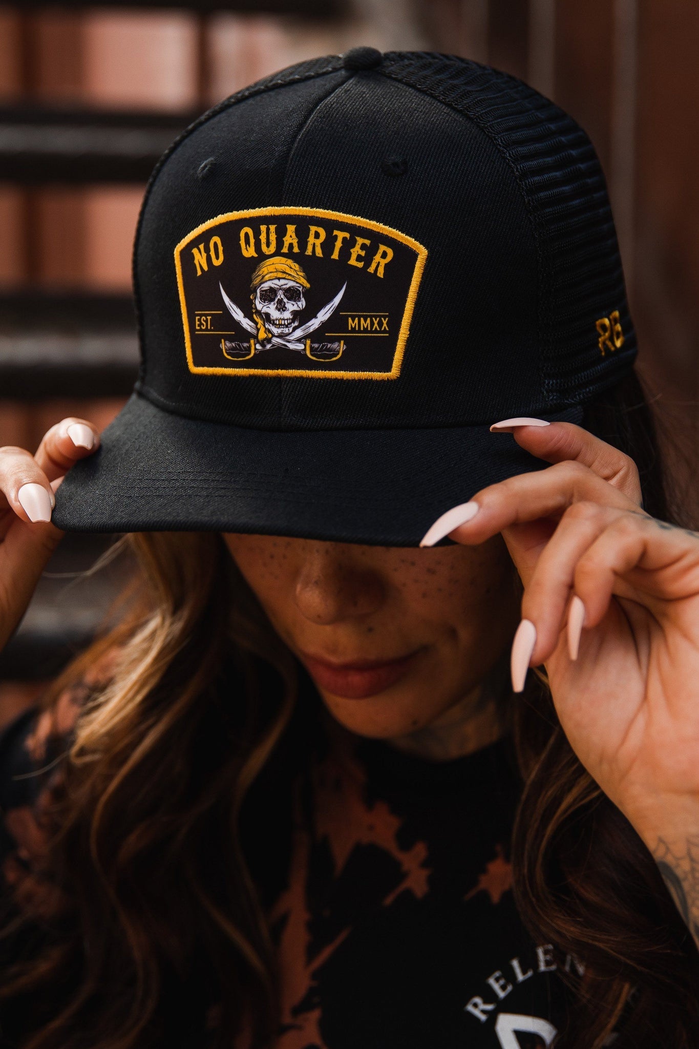 A person with long hair adjusts the Relentless Betrayal No Quarter Black Trucker Snapback, which features a yellow skull-and-crossbones NO QUARTER patch. Wearing a black shirt and sporting long, painted nails, they achieve the perfect fit with its snapback enclosure as the background stays blurred.