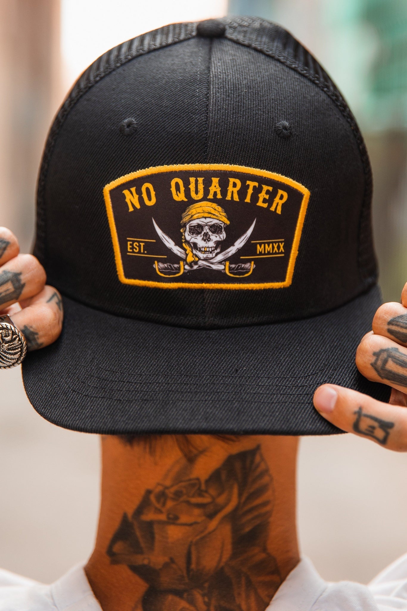A person adjusts their No Quarter Black Trucker Snapback by Relentless Betrayal, showcasing a skull and crossed knives logo with No Quarter Est. MMXX while revealing a rose tattoo on their neck.
