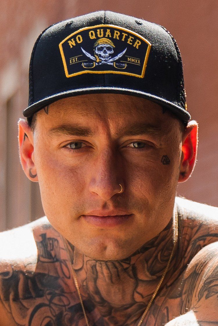 A man in a No Quarter Black Trucker Snapback by Relentless Betrayal, featuring a skull motif and snapback closure, stares ahead. His facial tattoos, nose ring, and tattooed chest create a striking image against the blurred outdoor backdrop.