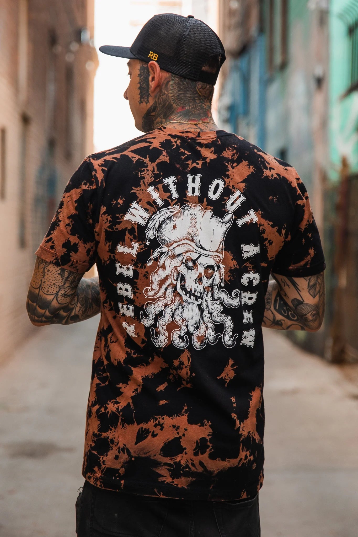 A tattooed rebel in a cap and a Relentless Betrayals Rebel Without A Crew Bleach Wash Tee with an octopus pirate design stands alone in an alleyway, embodying the outliers essence, as viewed from behind.