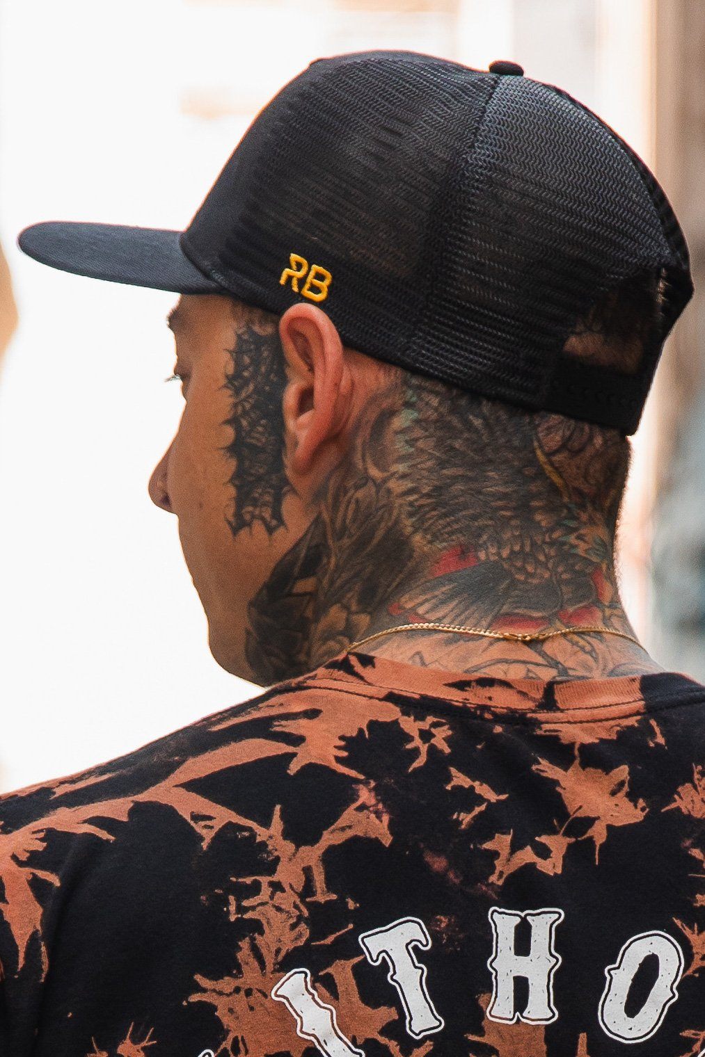 A person sporting a Relentless Betrayal No Quarter Black Trucker Snapback and a tie-dye shirt, proudly displaying neck and head tattoos. The shirt features partially visible white text on the back.