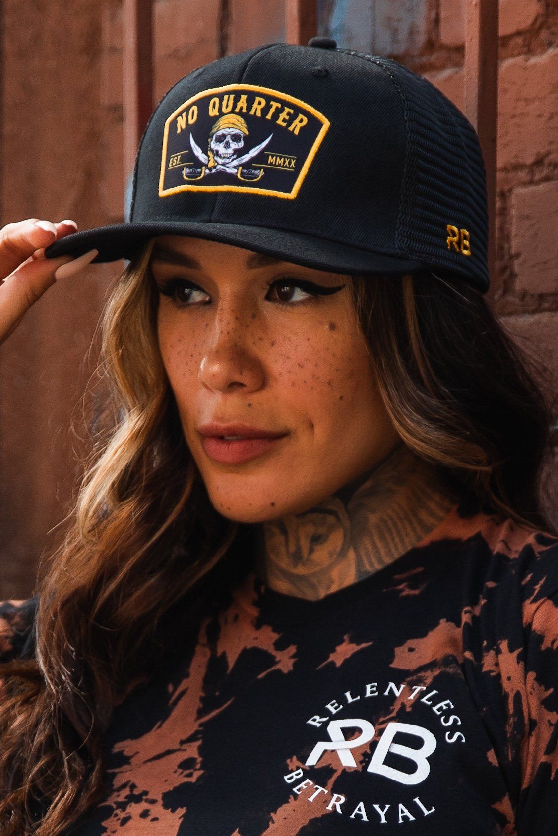 A person with long wavy hair and freckles wears a No Quarter Black Trucker Snapback, featuring a skull design and No Quarter text. Their black and orange tie-dye shirt from Relentless Betrayal reads the same phrase, complementing the edgy look against a brick wall.