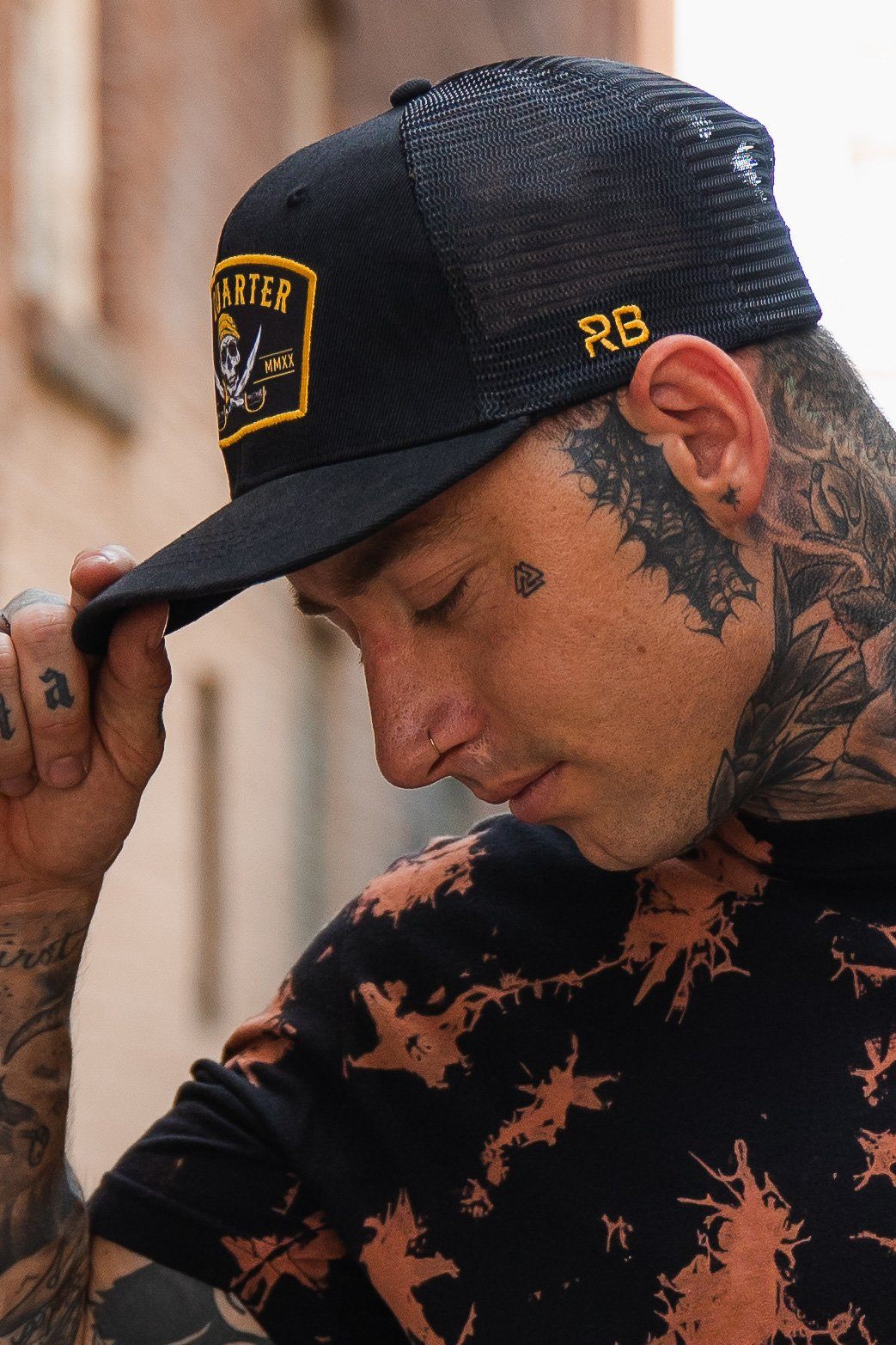 With tattoos on their neck and hand, a person adjusts the brim of a No Quarter Black Trucker Snapback by Relentless Betrayal, featuring yellow lettering. Their outfit includes a black and orange shirt in an outdoor setting.