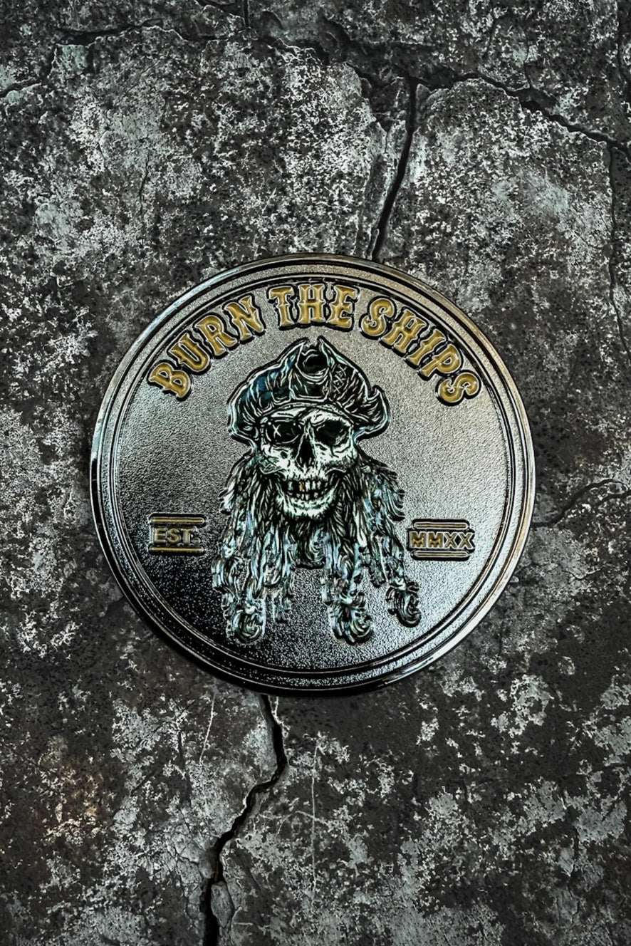 The Burn The Ships Challenge Coin by Relentless Betrayal is a circular emblem hand-forged from stainless steel, displaying a skull with a pirate hat and long hair. BURN THE SHIPS arches above, and EST MMXX sits below on a textured, cracked surface evoking rugged piracy.