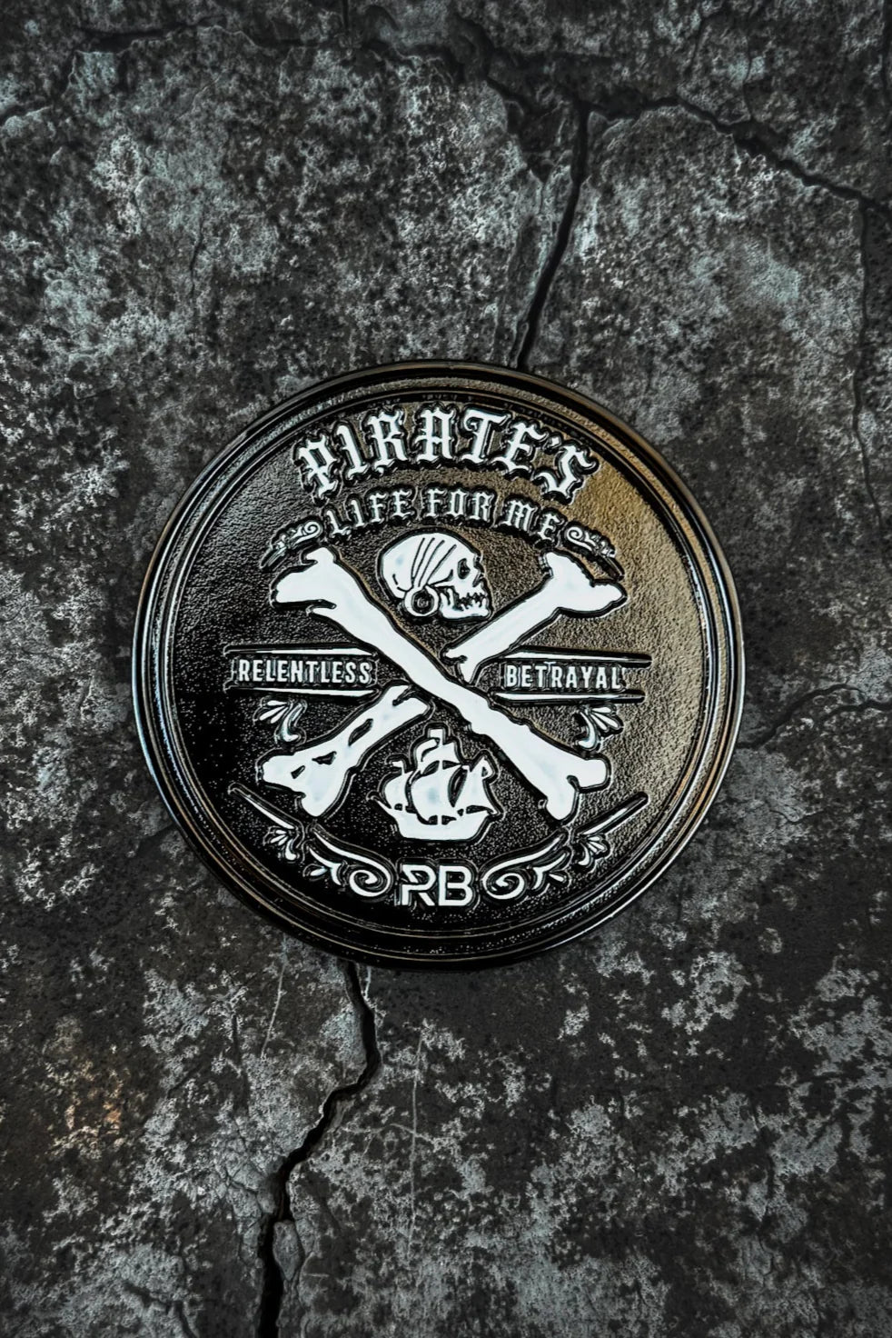 A handcrafted round emblem on a cracked, rocky surface features a pirate skull, crossbones with an anchor, and the text Pirates, Life for Me. It celebrates Relentless Adventure and displays initials PB, representing the Pirates Life Challenge Coin by Relentless Betrayal.