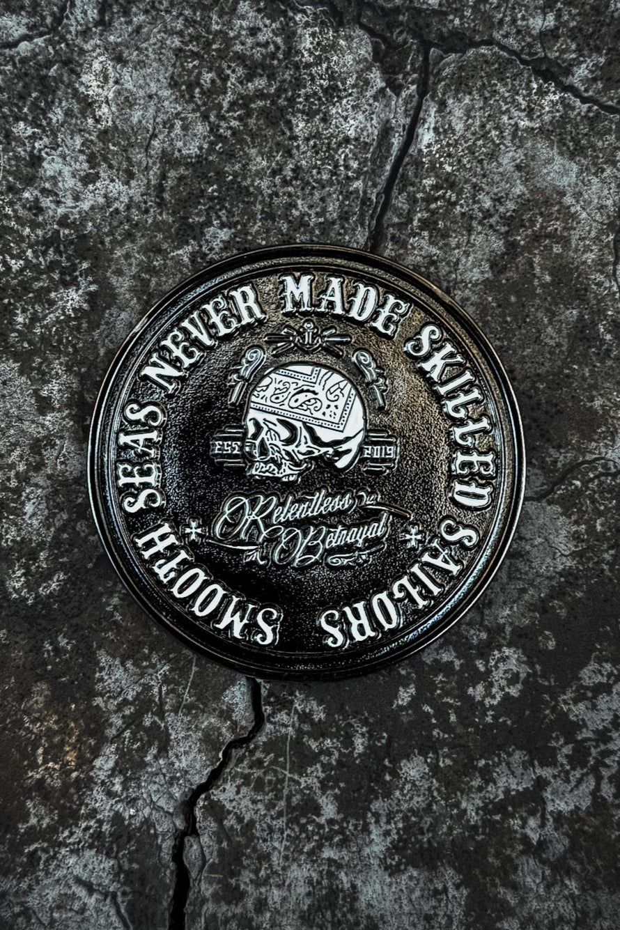 The Skilled Sailor Challenge Coin by Relentless Betrayal displays a round black and white design featuring a helmeted skull with the text Smooth seas never made skilled sailors, part of their resilience collection, shown on a rugged, cracked surface.