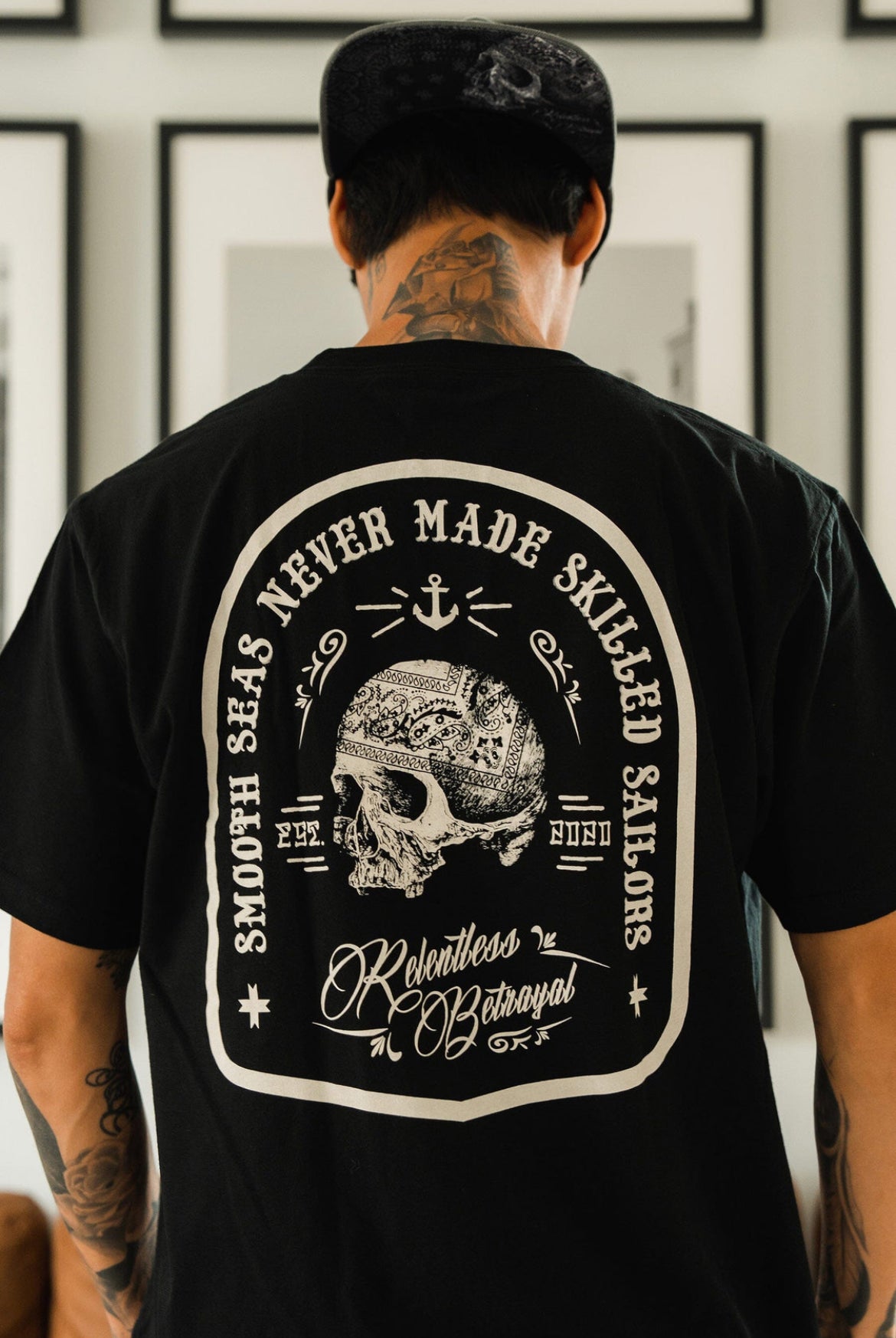 A tattooed man dons a black cap and the Skilled Sailor Tee by Relentless Betrayal, featuring a skull with a helmet, an anchor above, and the phrase Smooth Seas Never Made Skilled Sailors, symbolizing resilience and overcoming challenges.
