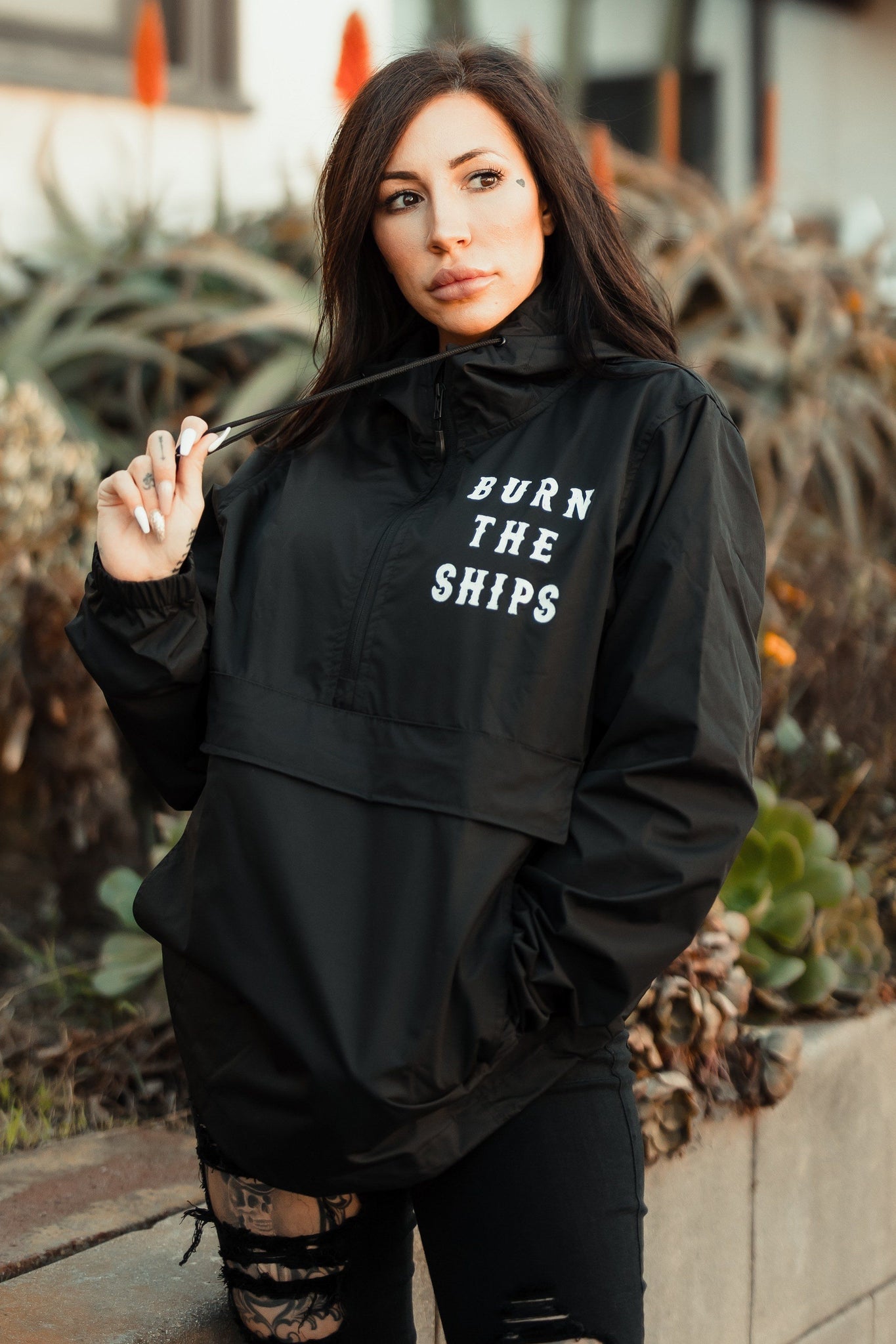 A person with long dark hair stands outdoors wearing the Burn the Ships Pullover Rain Jacket by Relentless Betrayal. This unisex, water-resistant jacket features BURN THE SHIPS on the front. They hold the collar with one hand, surrounded by lush plants.