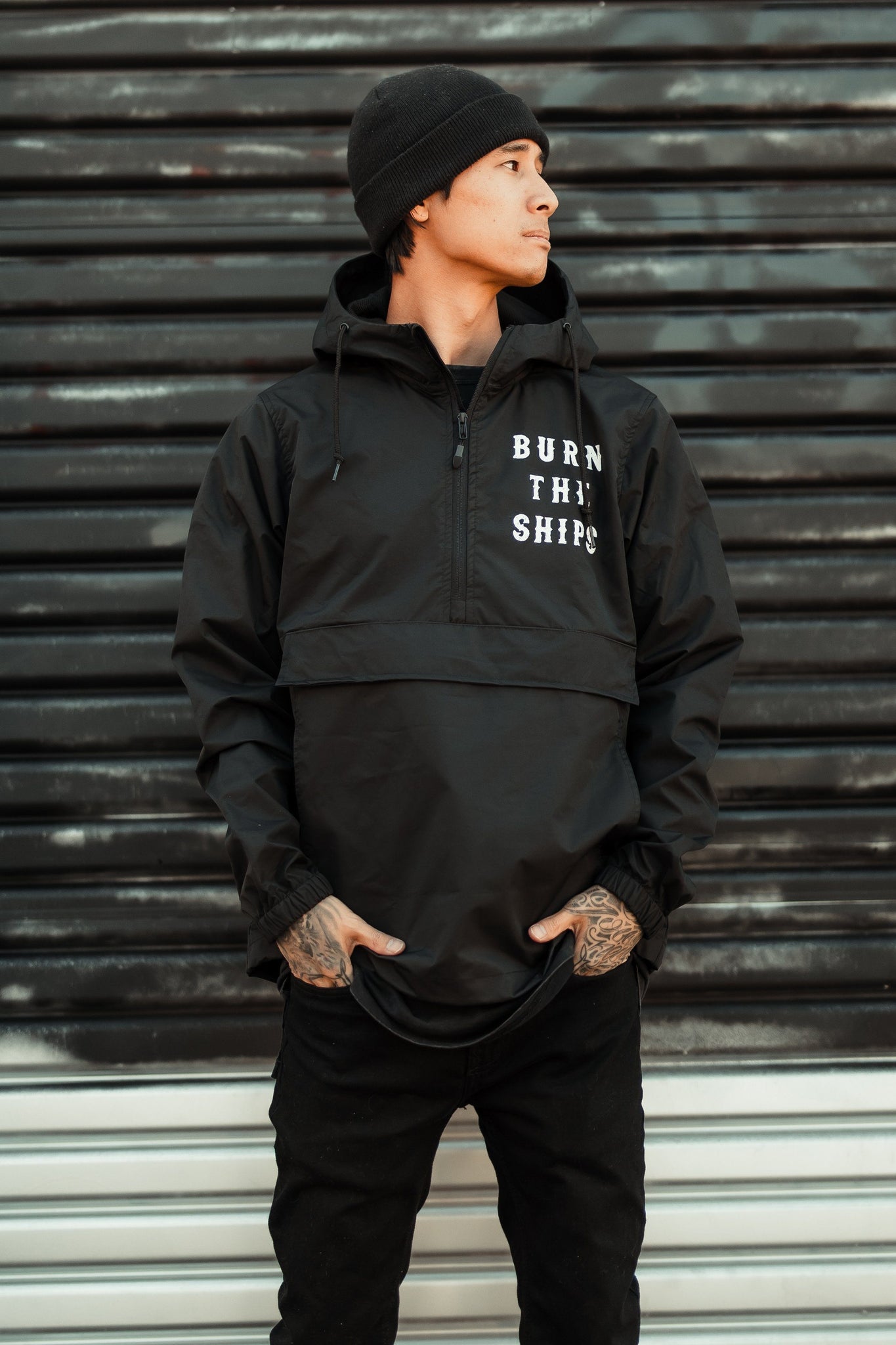A person in a beanie and hooded jacket stands against a corrugated metal backdrop, looking to the side. The Relentless Betrayal Burn the Ships Pullover Rain Jacket features BURN THE SHIPS on the front, has a water-resistant coating, and pockets for casually tucked hands.