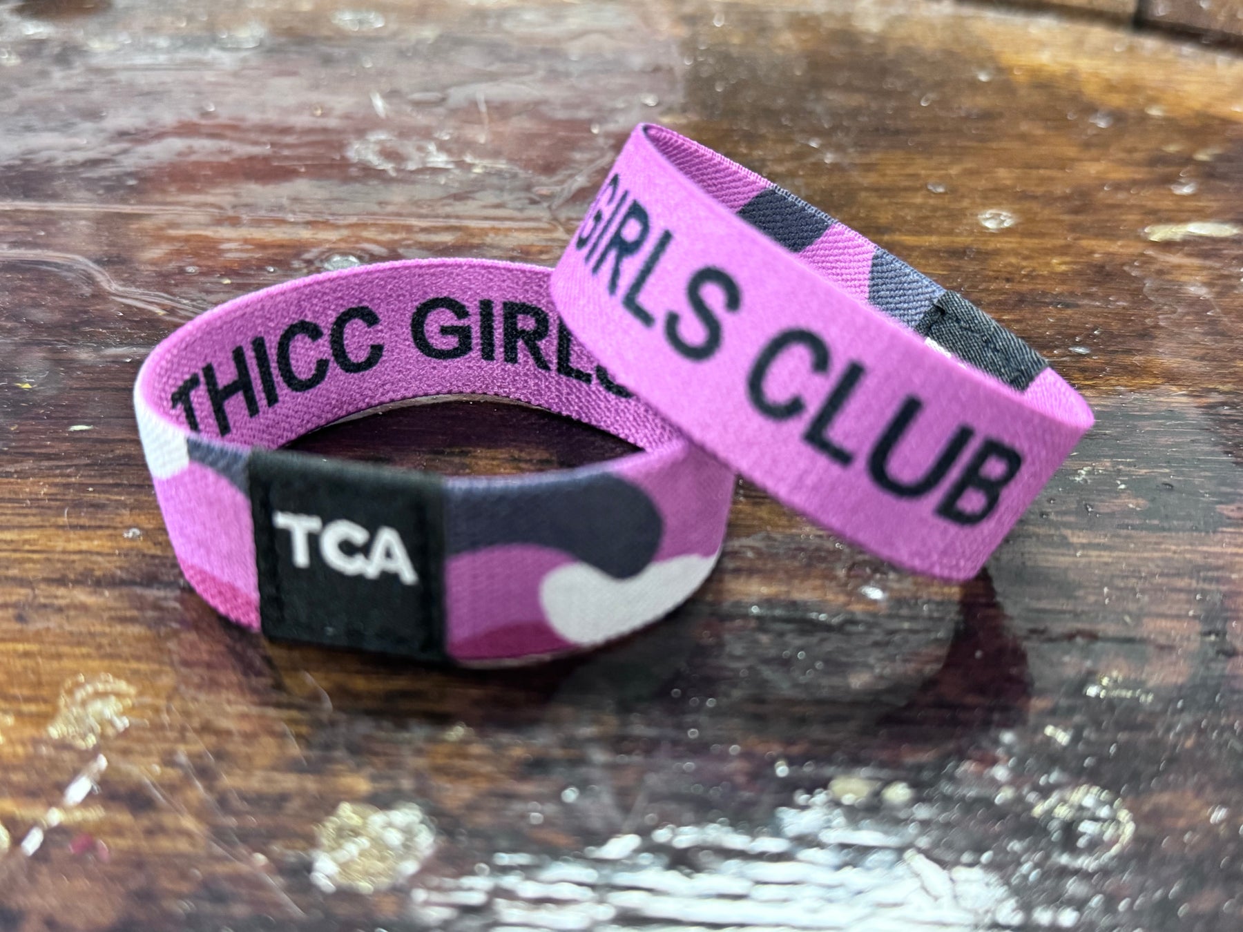 Two purple wristbands from Tango Charlie Apparels Thicc Girls Club rest on wood. One is upright, showing THICC GIRLS CLUB in a black camouflage pattern, symbolizing body positivity. The other lies flat with TCA in white on black—ideal for fans of empowering ladies’ apparel.