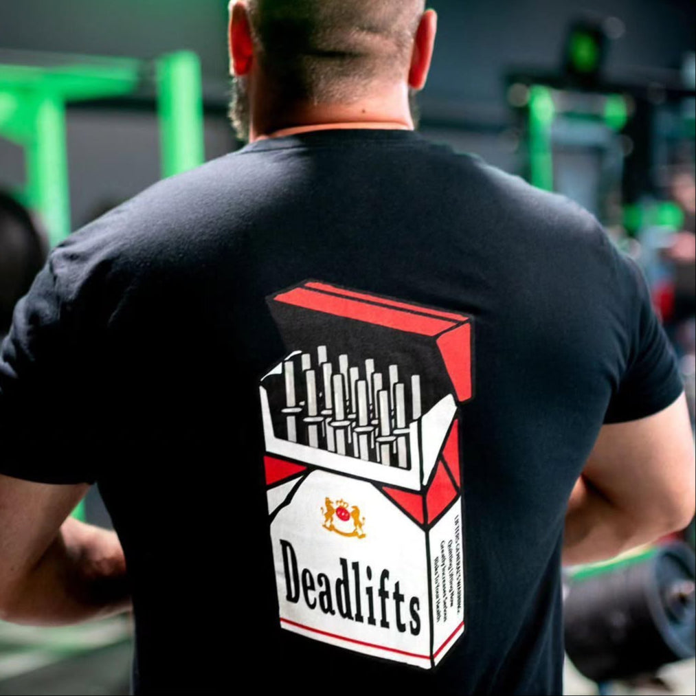 In a gym, a man wears the Deadlift Addiction - Tee by Tango Charlie Apparel. The black shirt flaunts a cigarette pack graphic labeled Deadlifts, where barbell plates replace cigarettes. A lifters warning adds charm, with green gym equipment subtly visible in the background.