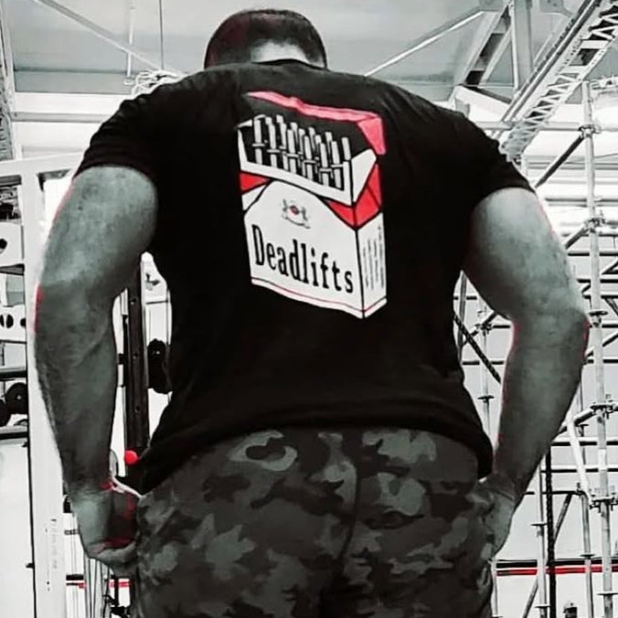 In the gym, a person wears a Deadlift Addiction - Tee from Tango Charlie Apparel, echoing once you start deadlifting, you cant stop, with camo shorts and muscular arms completing their look.