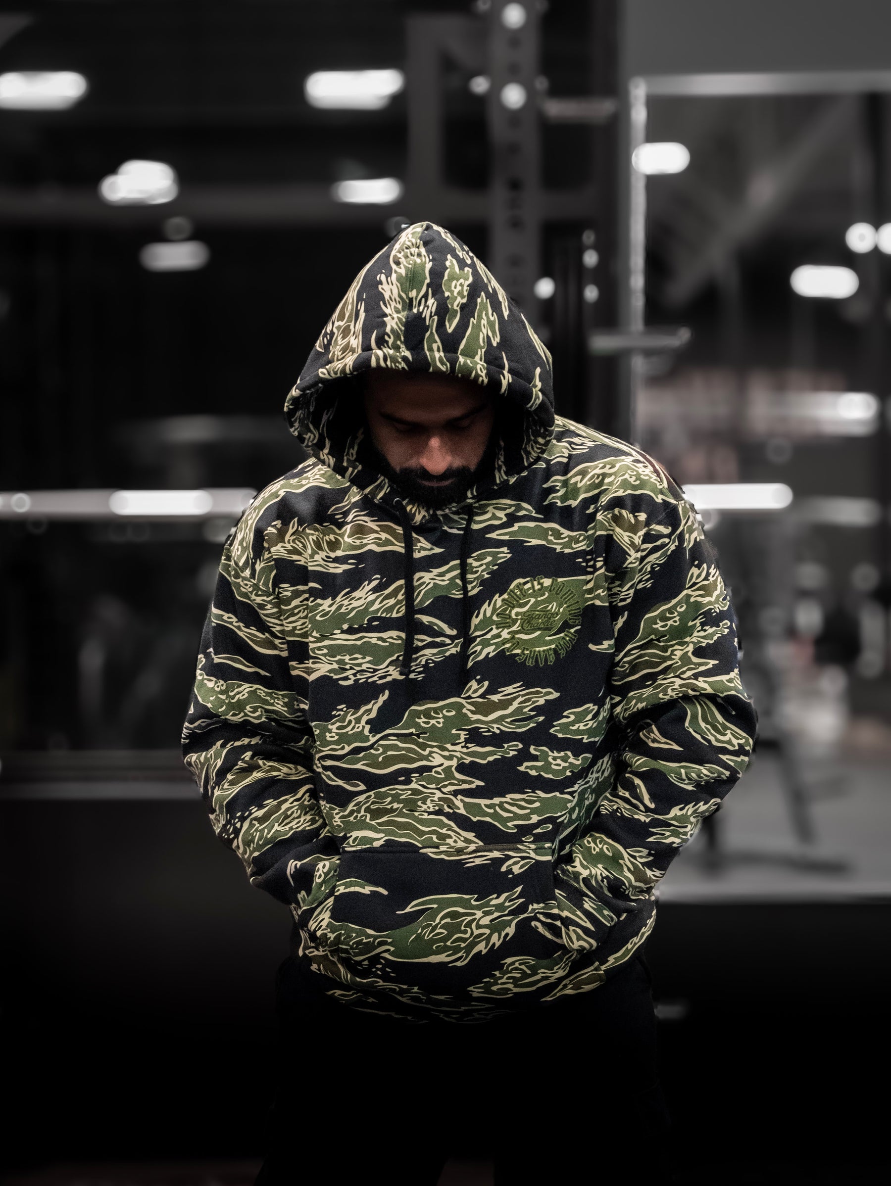 A person stands with head down and hands in pockets, wearing the No One Is Coming To Save You hoodie by Tango Charlie Apparel. The dimly lit background suggests a gym, highlighting their focus on self-reliance and personal goals.