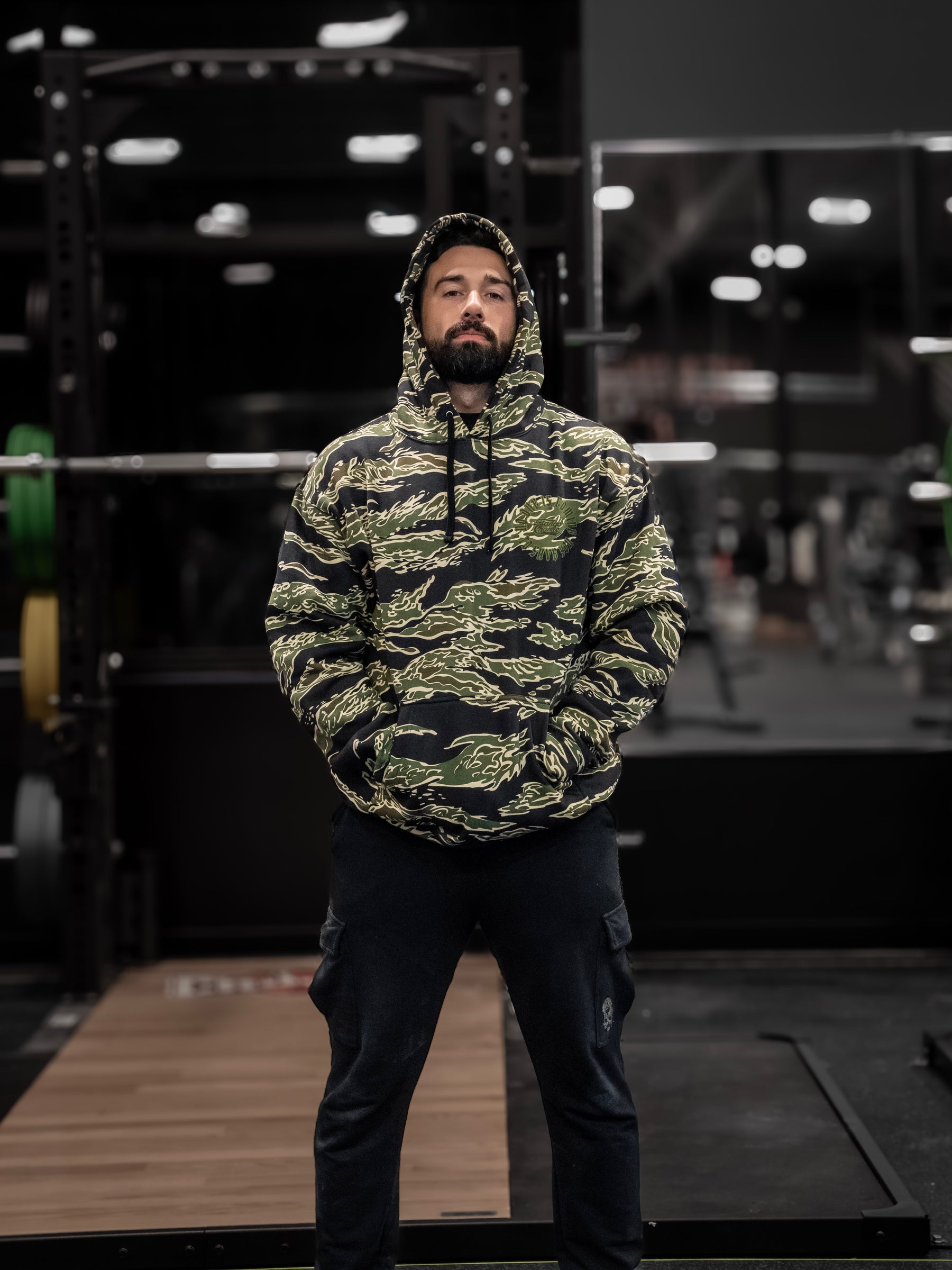 In the gym, a person stands confidently wearing the No One Is Coming To Save You hoodie by Tango Charlie Apparel. With hands in their pocket and gym equipment blurred behind them, they embody self-reliance and focus on personal accountability amid life goals.