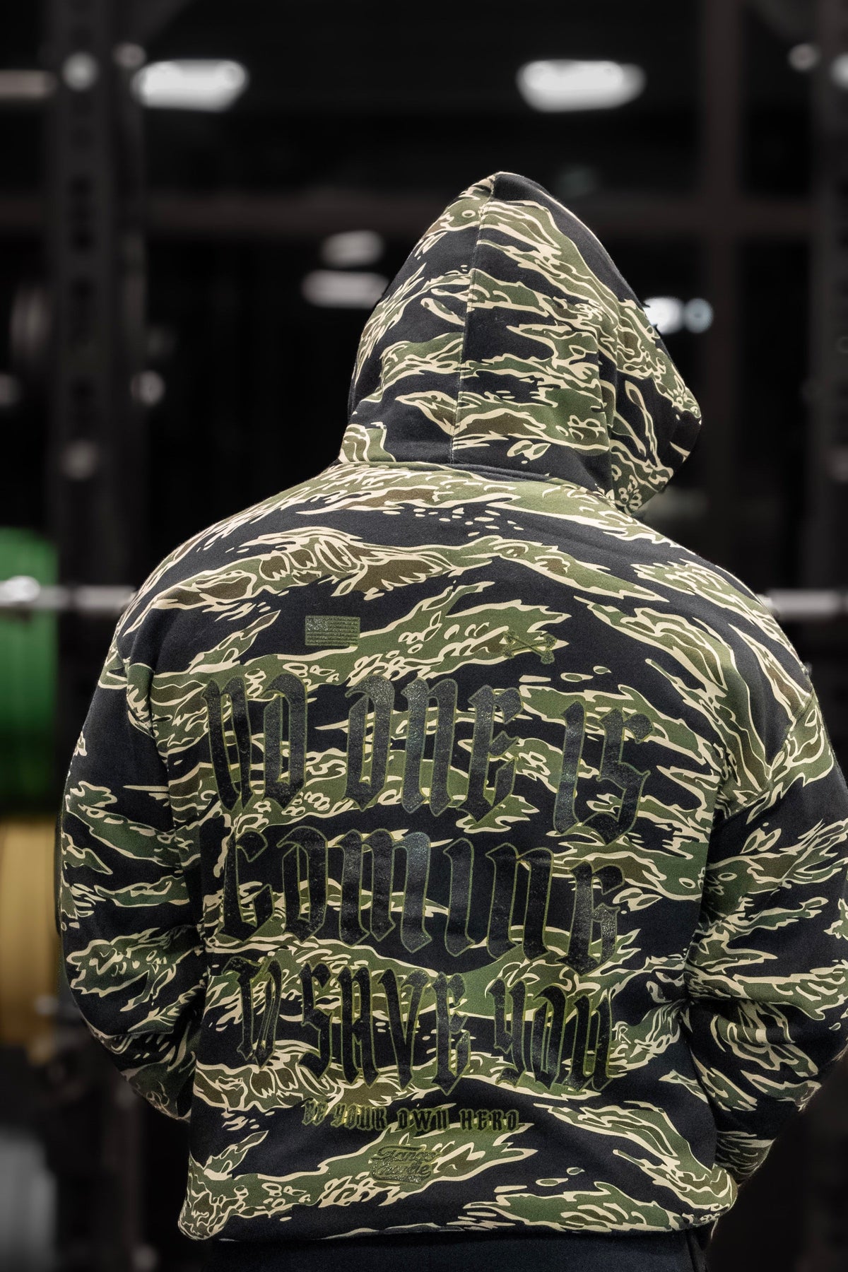 Wearing the "No One Is Coming To Save You" Premium Hoodie by Tango Charlie Apparel, a person stands with their back to the camera in front of a weight rack with green and yellow plates, embodying self-motivation at the gym.