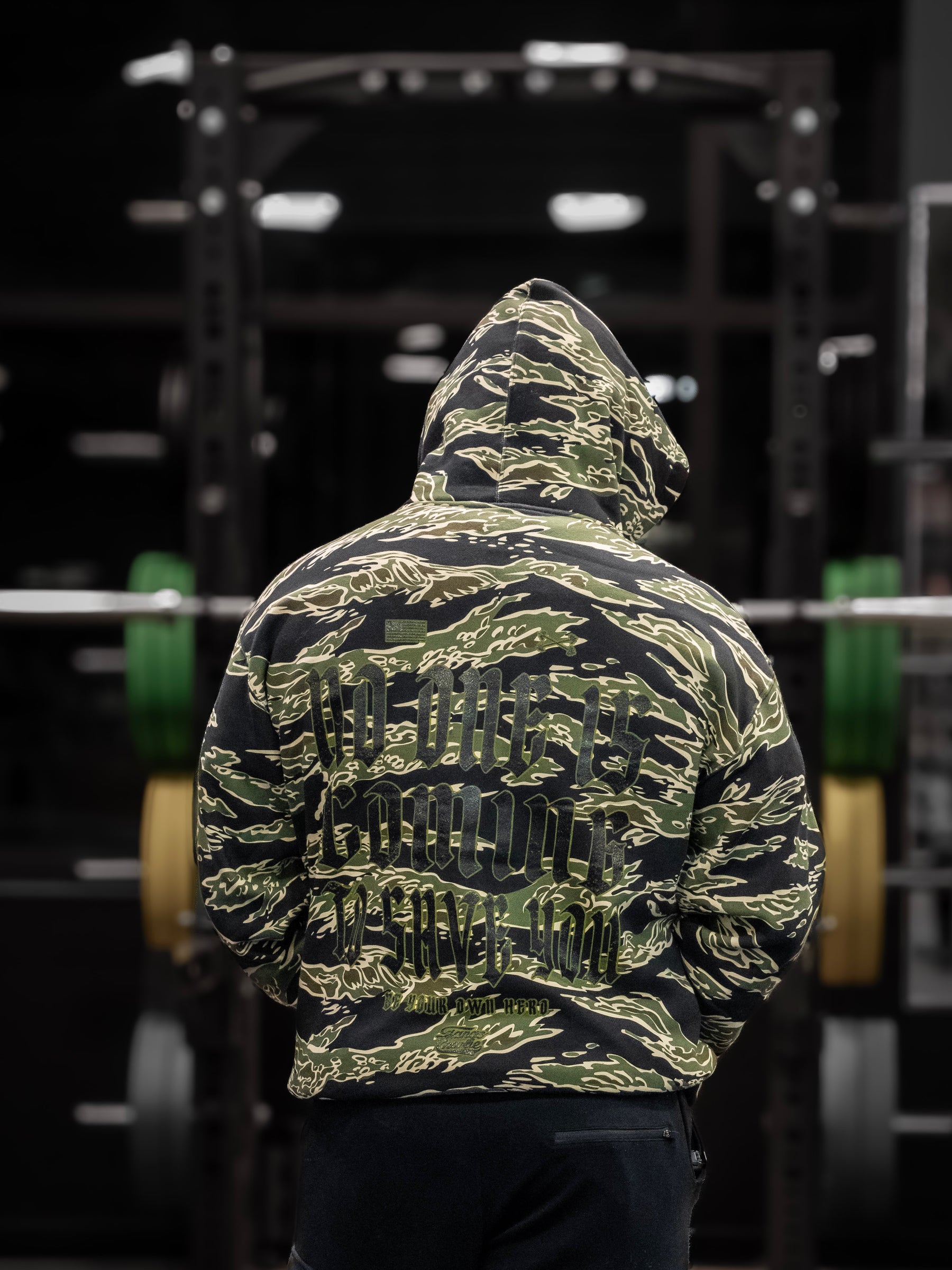 In a dim gym, someone dons the Tango Charlie Apparels No One Is Coming To Save You green and black camo hoodie, facing a barbell with green and yellow plates as they embrace self-reliance and determination in pursuit of their life goals.