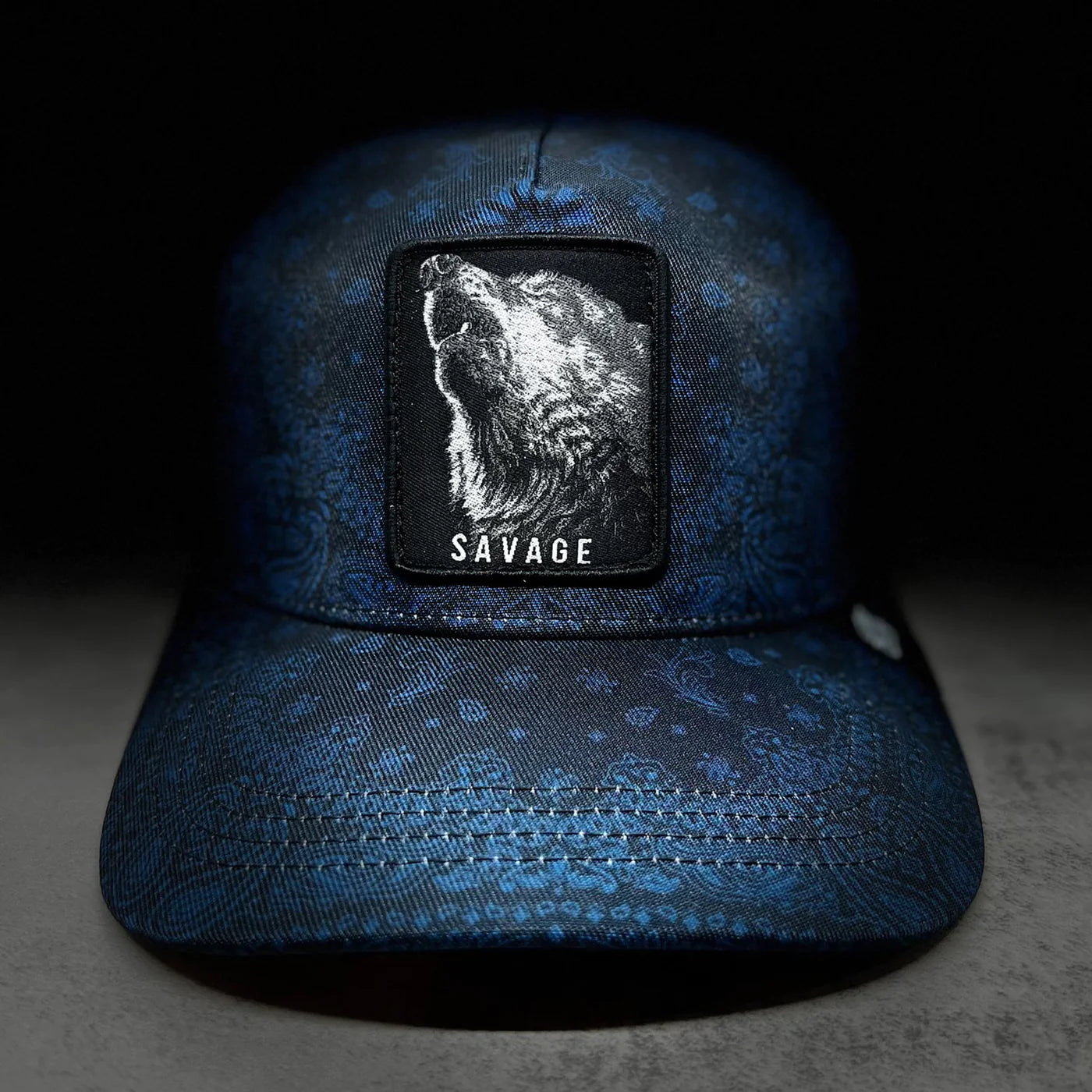 The Savage Slate Paisley-Trucker Snapback by Tango Charlie Apparel is a blue hat featuring a bear head patch with SAVAGE underneath, highlighted on a dark background with textured paisley detail and a high-quality snapback enclosure.