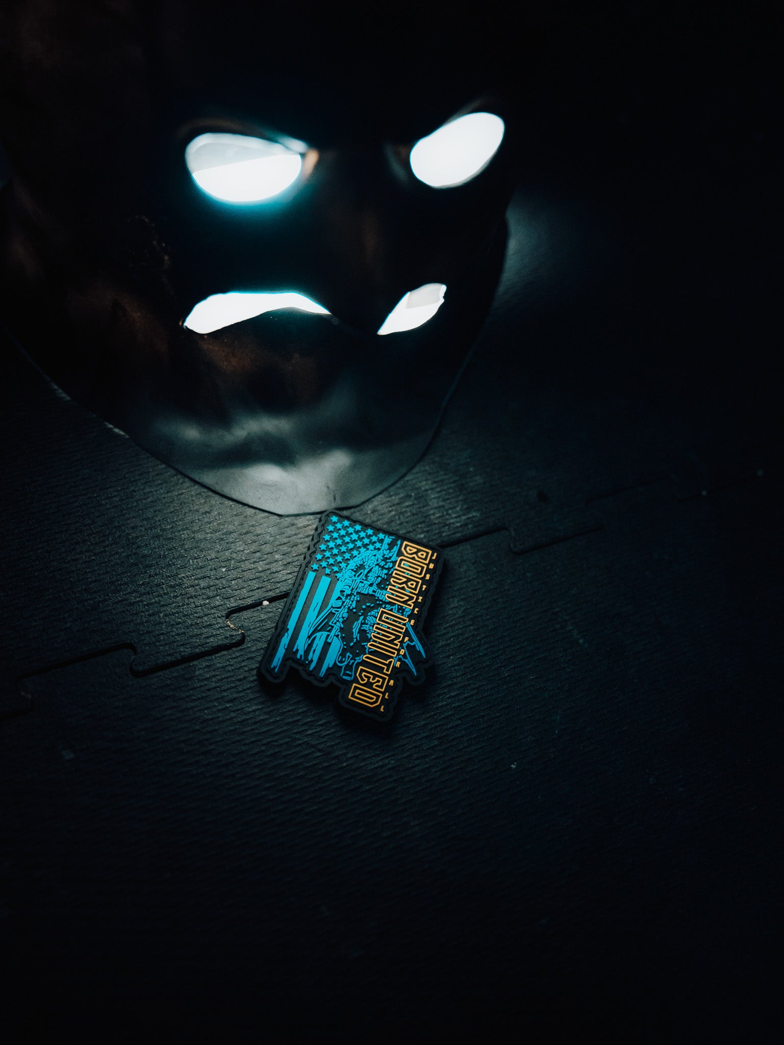 A mask with glowing eyes lies on a dark surface, illuminating a vibrant pin with Comic-Con and an illustrated skyline. Beside it, the Justice For All PVC Patch from Born United Private Label adds flair, echoing the scenes vivid hues.