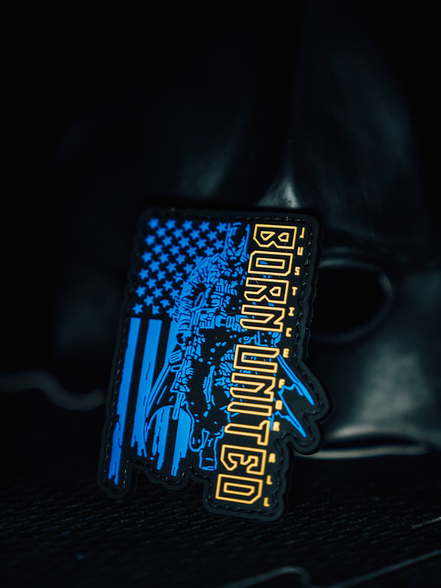 The Justice For All PVC Patch by Born United Private Label is a waterproof, washable patch with an Arizona shape featuring the American flag, a skull design, and BORDER UNITED text in blue and yellow against a dark background. Its crafted with hook & loop for easy attachment.