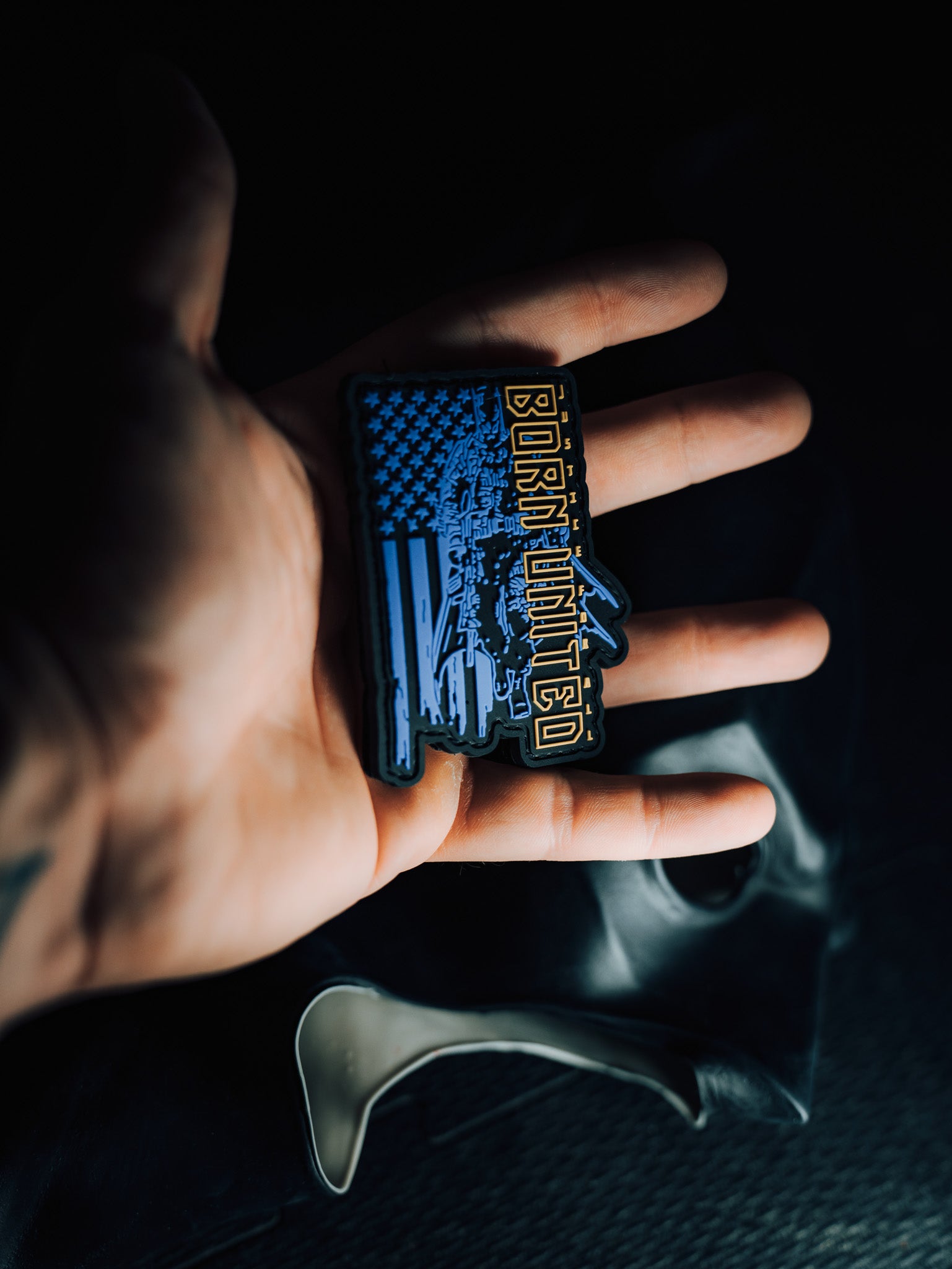 A hand displays the Justice For All PVC Patch by Born United Private Label, featuring bold Born United text. Resembling an American Flag with stars and abstract shapes, this waterproof, washable patch symbolizes unity and resilience against a dark backdrop.
