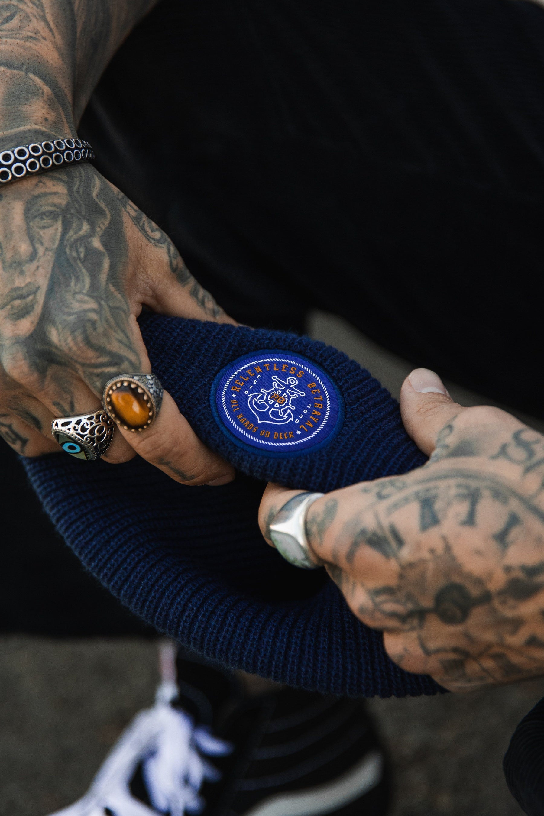 Tattooed hands clutch a navy All Hands On Deck Beanie from Relentless Betrayal, featuring an anchor patch. The person wears multiple rings and a beaded bracelet, with black and white sneakers also visible.
