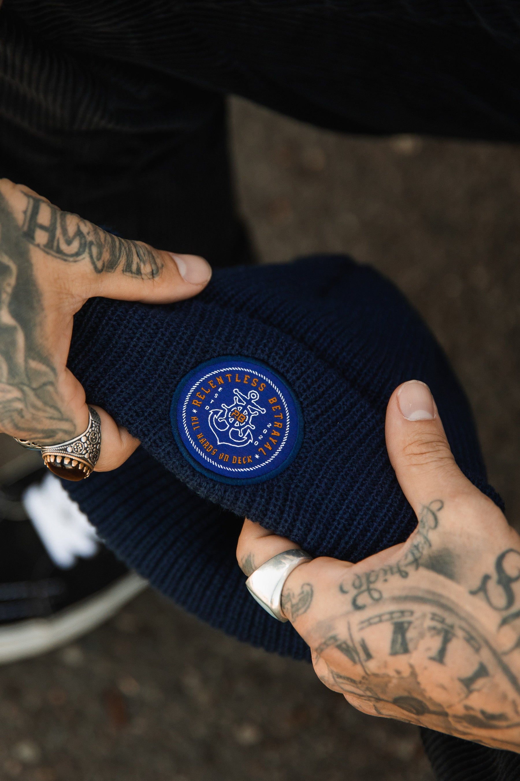 Tattooed hands adjust a navy blue All Hands On Deck Beanie, featuring a vintage-style Relentless Betrayal logo patch. Gold rings complement the tattoos against a blurred background.