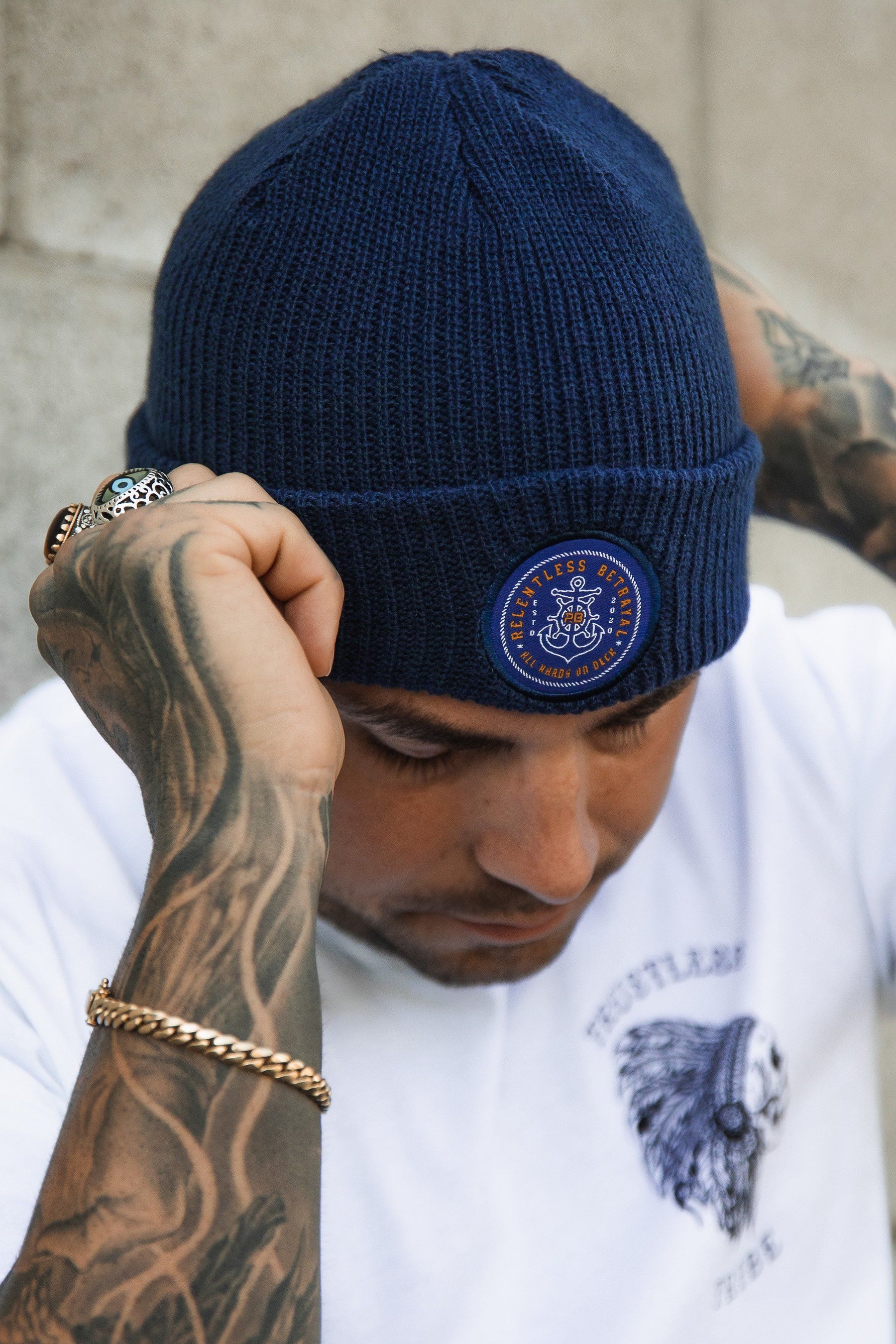 A tattooed person adjusts their All Hands On Deck Beanie by Relentless Betrayal, wearing a logoed white T-shirt featuring an eagle. A chain bracelet with gold details and a decorative ring adorn the scene against a textured gray wall.