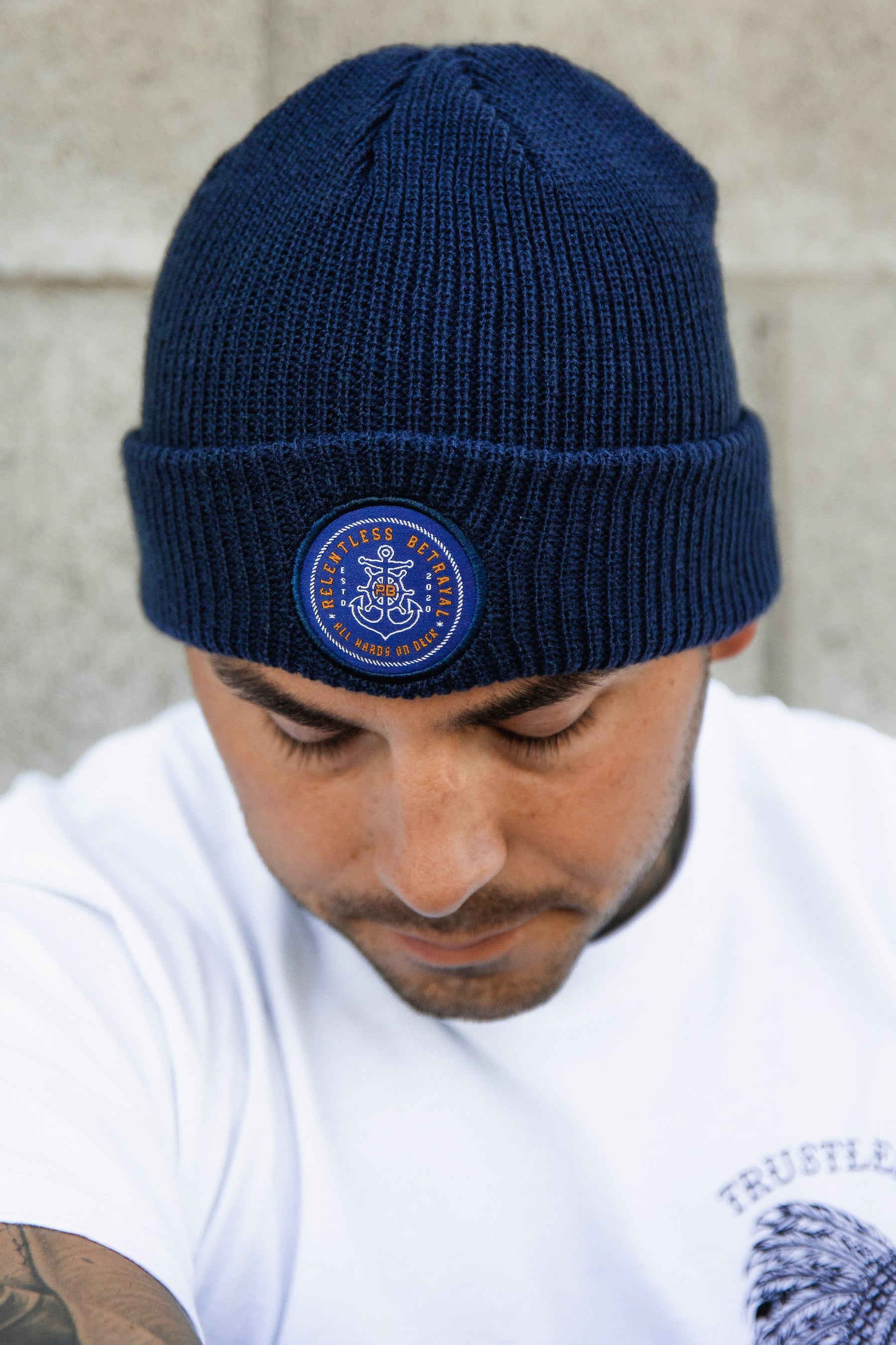 A person in a dark blue acrylic All Hands On Deck Beanie by Relentless Betrayal, featuring a circular fearless patch and anchor, glances down while wearing a lion graphic white T-shirt against a concrete wall backdrop.