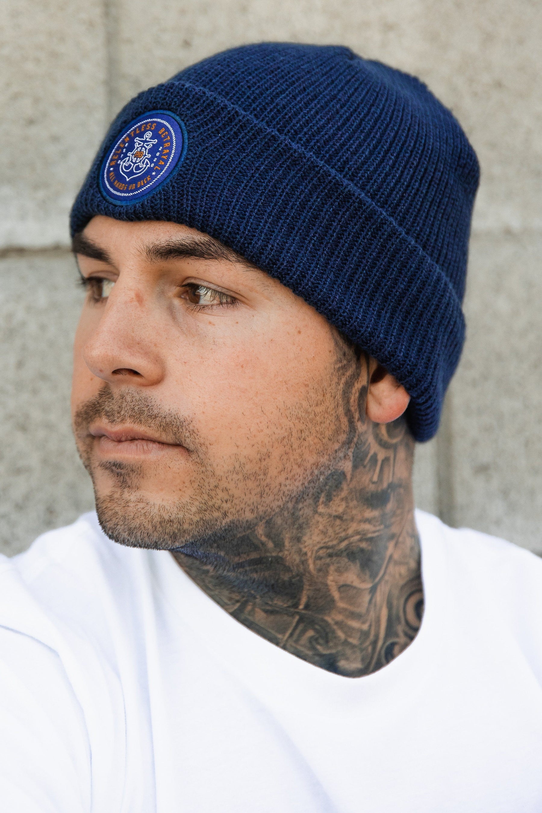 A person with a tattooed neck wears a Relentless Betrayals All Hands On Deck Beanie with an embroidered logo. Gazing to the side in a white shirt, they stand against a gray brick wall.