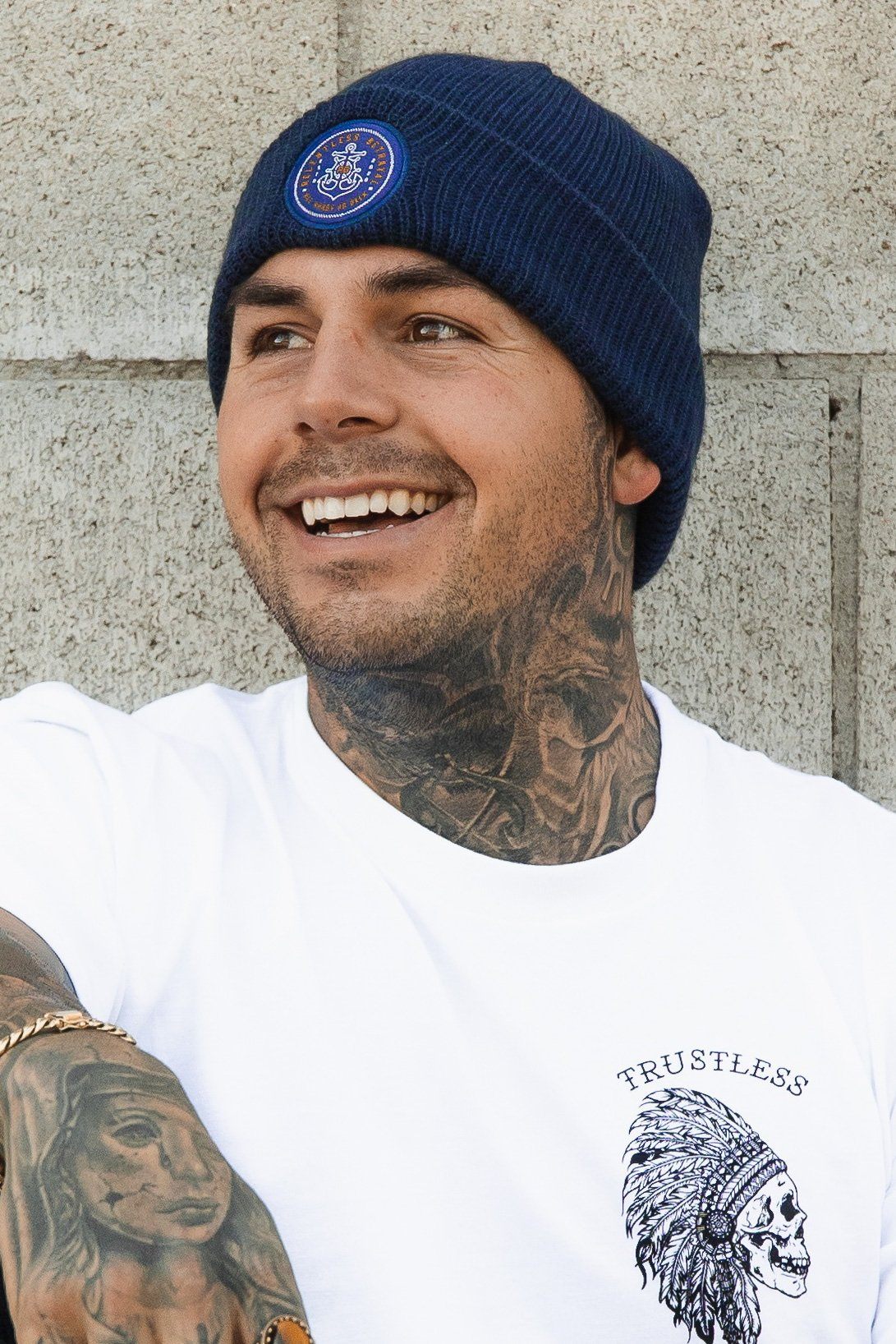 A man with tattoos, smiling in front of a textured concrete wall, wears a Relentless Betrayal All Hands On Deck Beanie in navy and a white T-shirt with an illustrated chest design.