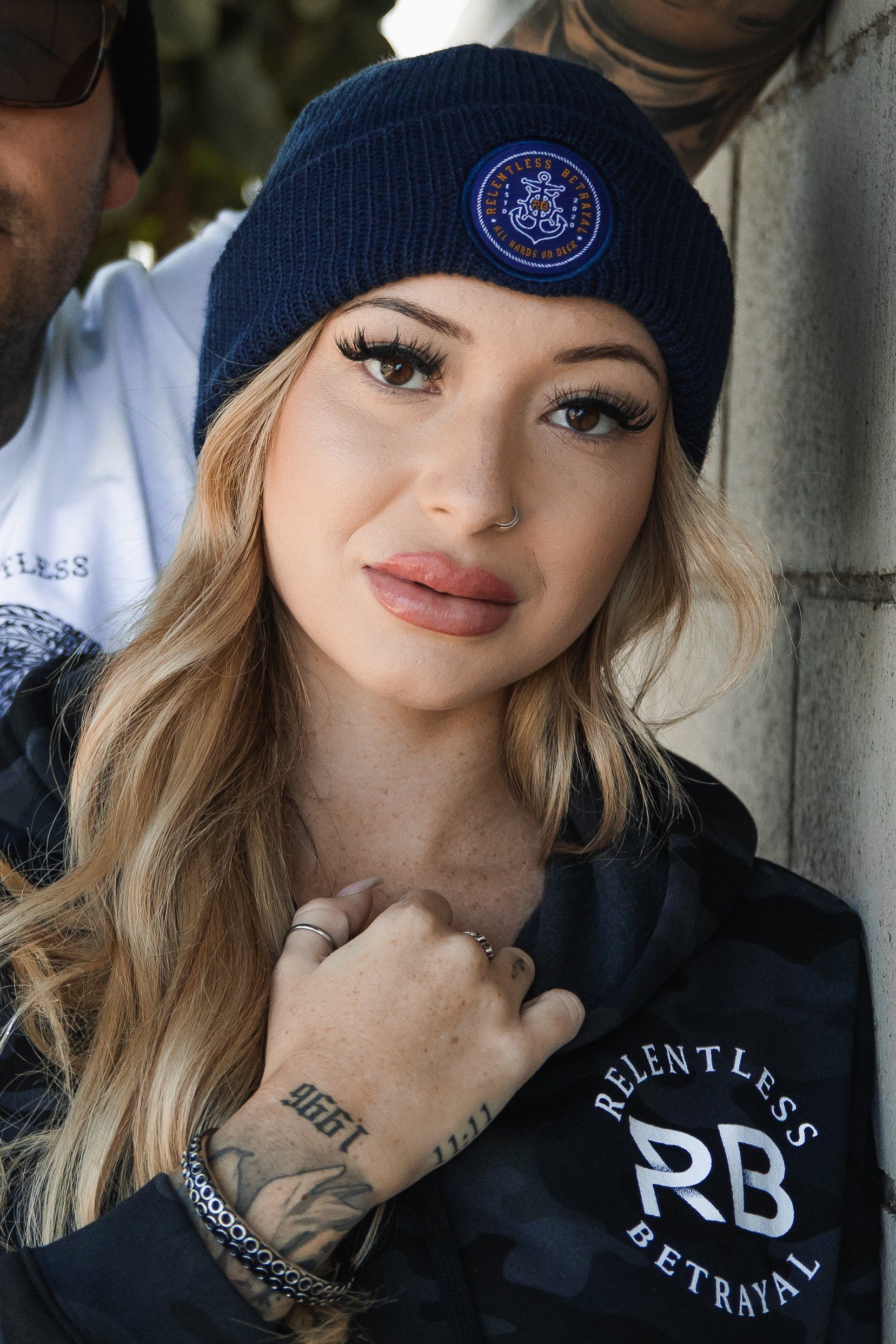 A long-haired blonde woman with a nose ring wears the All Hands On Deck blue acrylic beanie by Relentless Betrayal, paired with their dark camo jacket. Her tattooed hands and wrist complement her pose against a textured wall.