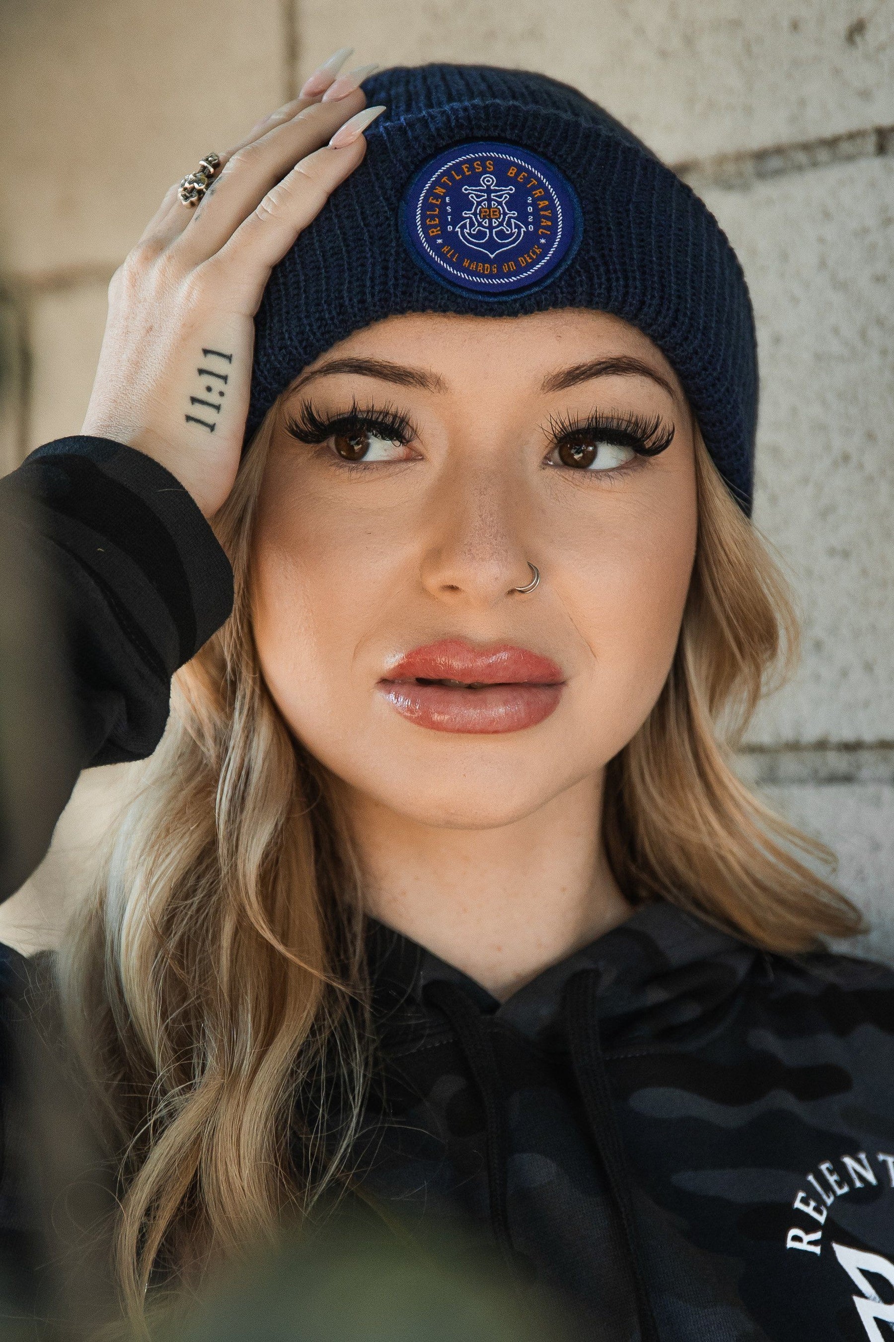 A person with long, wavy blonde hair and a hoop nose ring, sporting visible hand tattoos, gazes upward while wearing a black hoodie and holding a Relentless Betrayal All Hands On Deck Beanie with gold details against a textured wall.
