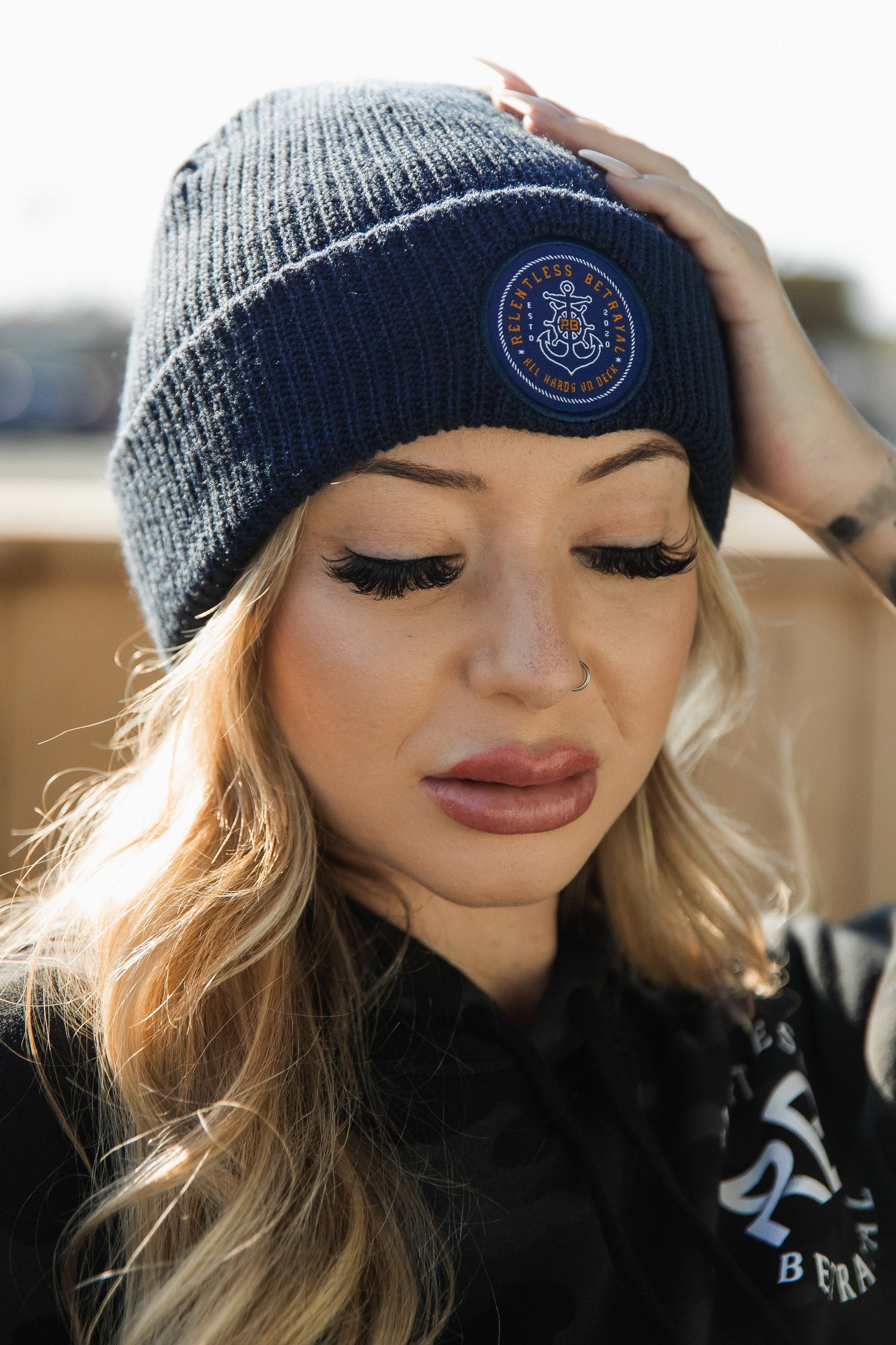 A person with long blonde hair wears a Relentless Betrayal All Hands On Deck Beanie with gold detailing and a black sweater. They have long eyelashes and look down, with a blurred background hinting at an outdoor location.
