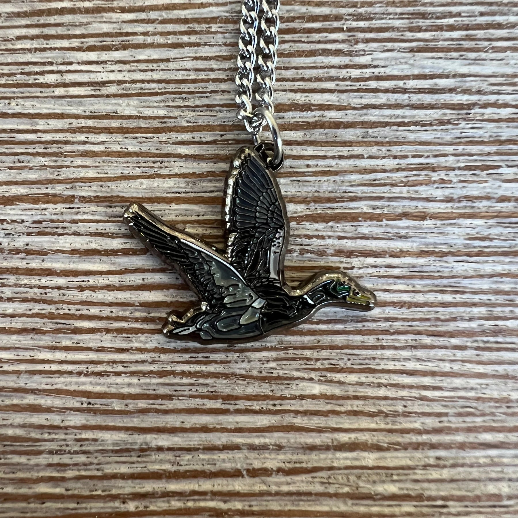 The Duck It OG Necklace by Mucker Bucket features a stainless steel flying bird pendant with detailed wings and body, set against a textured wood background and finished with a shiny surface. Proudly made in the USA.
