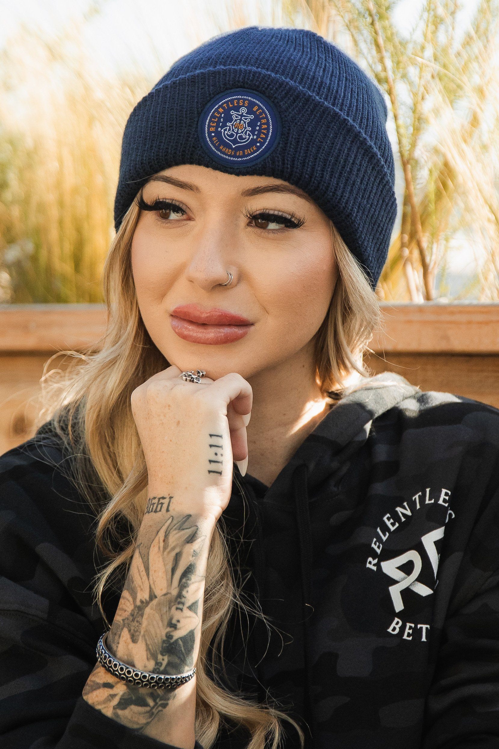 A woman with long blonde hair wears a Relentless Betrayal All Hands On Deck Beanie and a black hoodie as she poses thoughtfully outdoors, hand under chin. Tattoos decorate her hand and wrist, while tall grass and a wooden structure enhance the scene’s rustic charm.