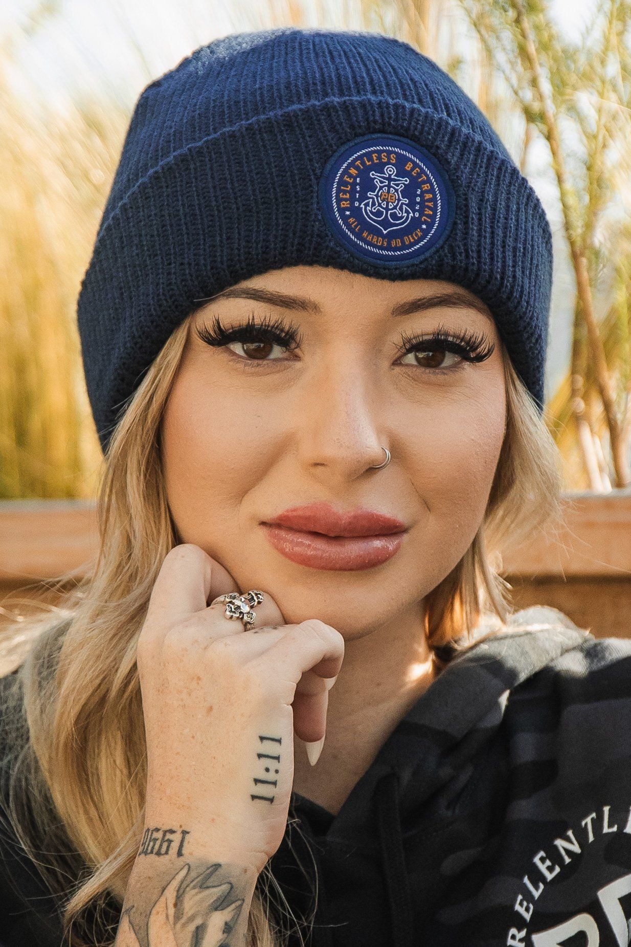 A person dons Relentless Betrayals All Hands On Deck Beanie, a navy blue acrylic piece with gold details, and a black hoodie. Their wrist and hand are tattooed, with long eyelashes accentuating their look. Tall grass and wooden elements complete the outdoor scene.