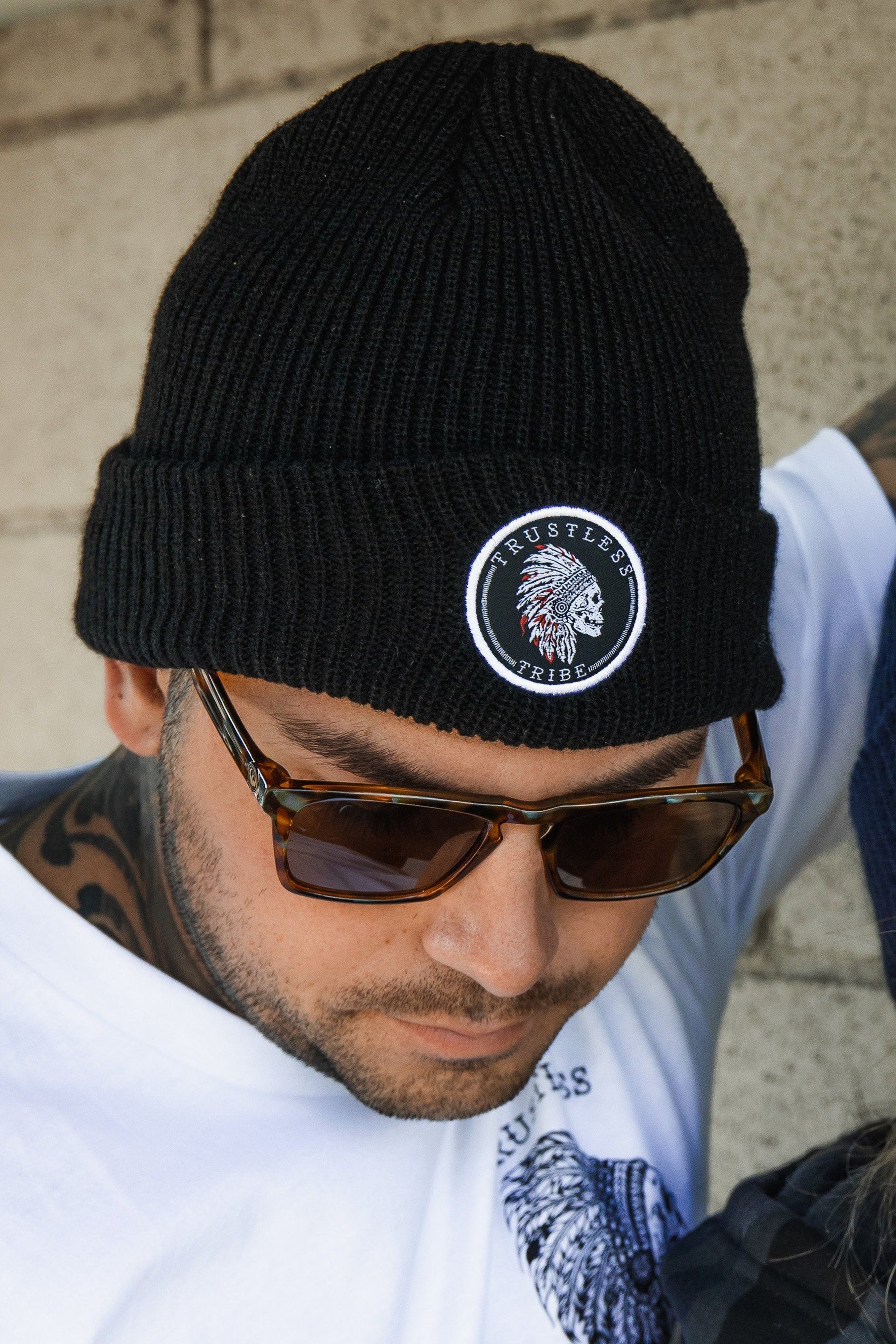 A person in a black acrylic Trustless Tribe Beanie by Relentless Betrayal, sunglasses, and a white graphic tee. They have a neck tattoo and stand against a beige wall, capturing the No Prey No Pay vibe.