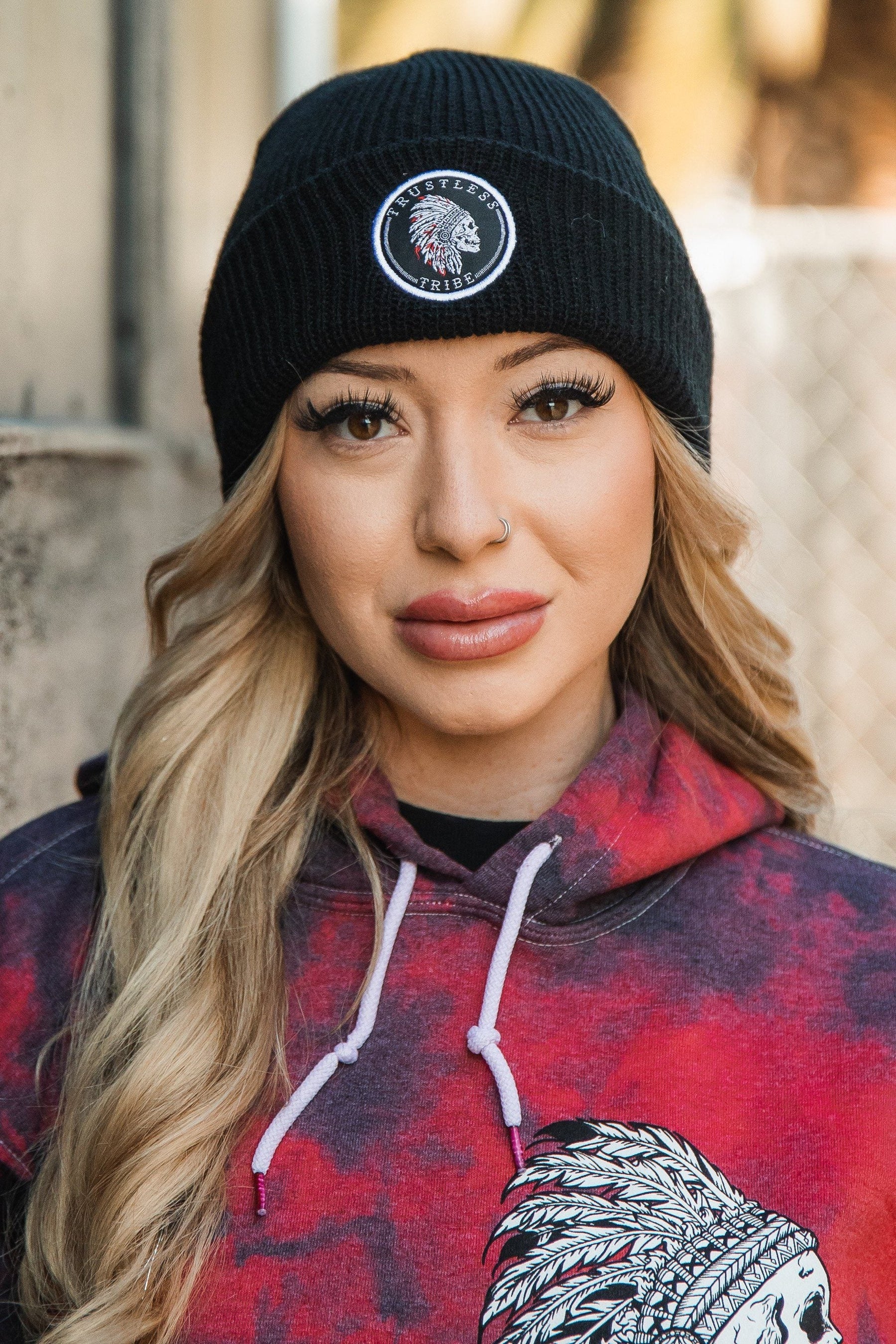 A blonde woman stands confidently outdoors wearing a Trustless Tribe Beanie by Relentless Betrayal and a red hoodie with a Native American design. Her look includes long eyelashes, light pink lipstick, and a nose ring, set against a fence and blurred scenery backdrop.