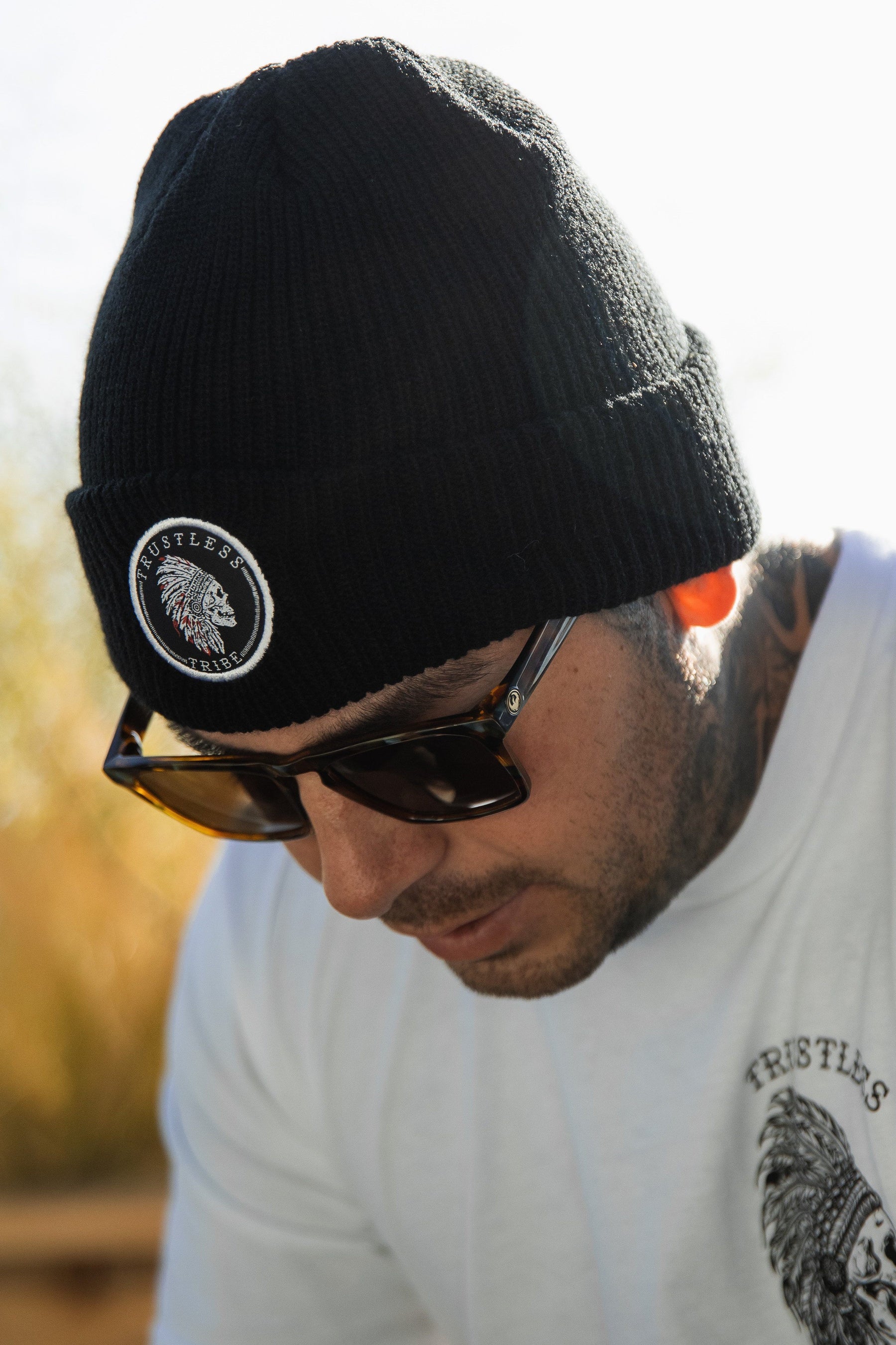 A person outdoors under a sunny sky wears a Trustless Tribe Beanie by Relentless Betrayal featuring a circular patch, sleek sunglasses, and a white T-shirt printed with No Prey No Pay.