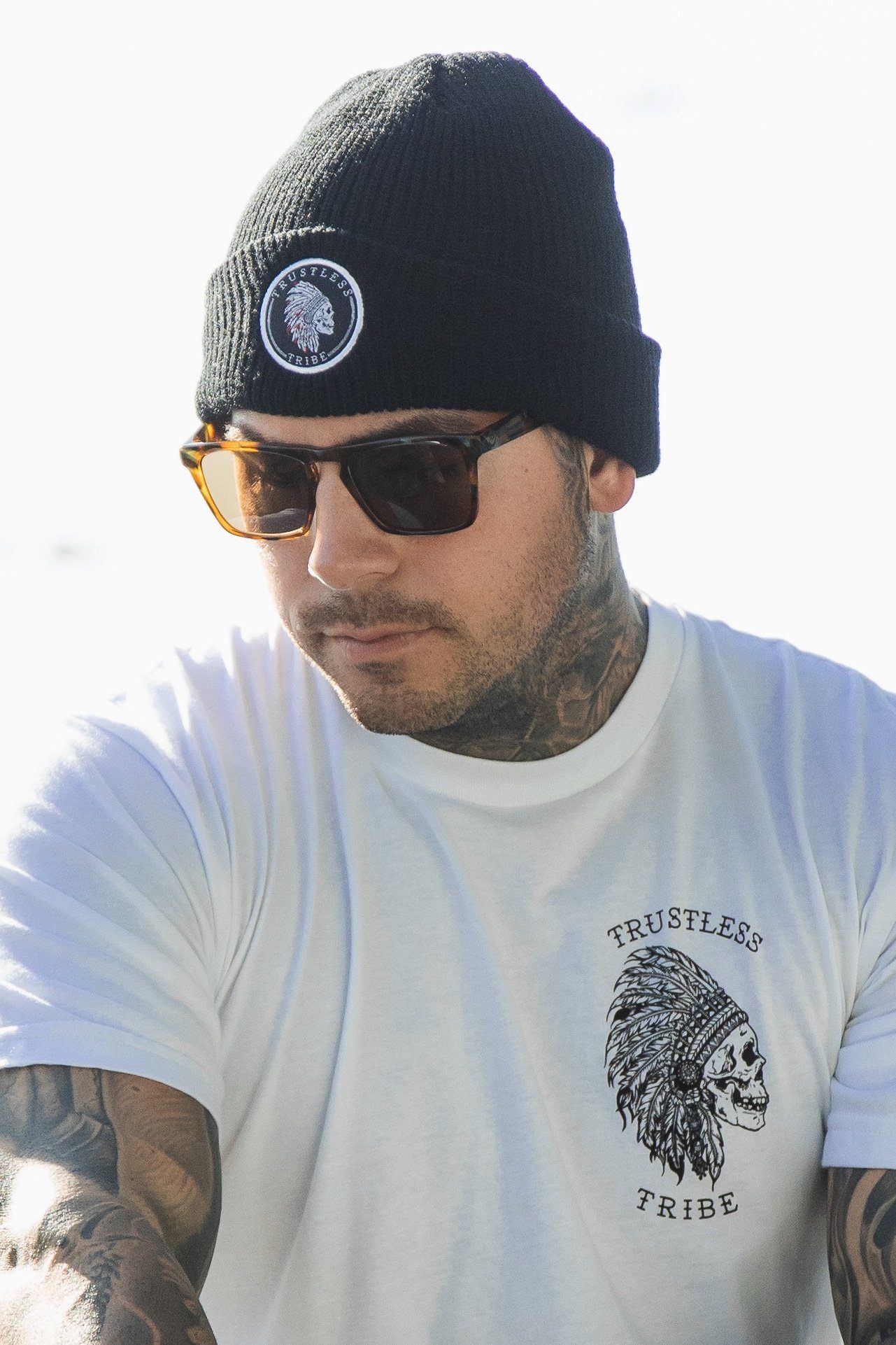 Wearing Relentless Betrayals sunglasses, a Trustless Tribe Beanie, and a white Trustless Tribe t-shirt with a skull in headdress, the tattooed individual gazes sideways.