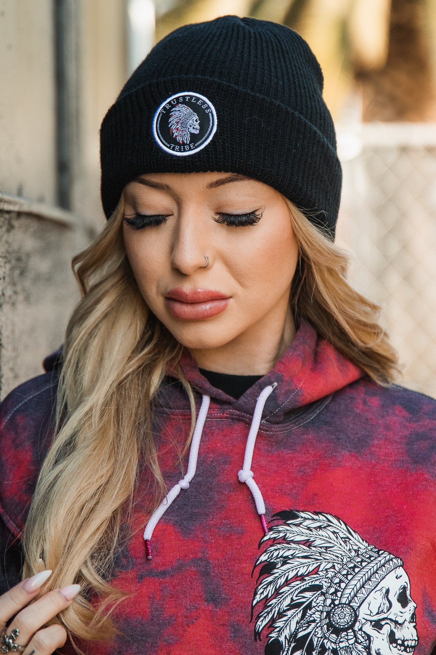 A person with long hair wears a Trustless Tribe Beanie by Relentless Betrayal and a red hoodie featuring a Native American chief design. They stand outdoors, near a fence and tree, looking down calmly, embodying the spirit of No Prey No Pay.
